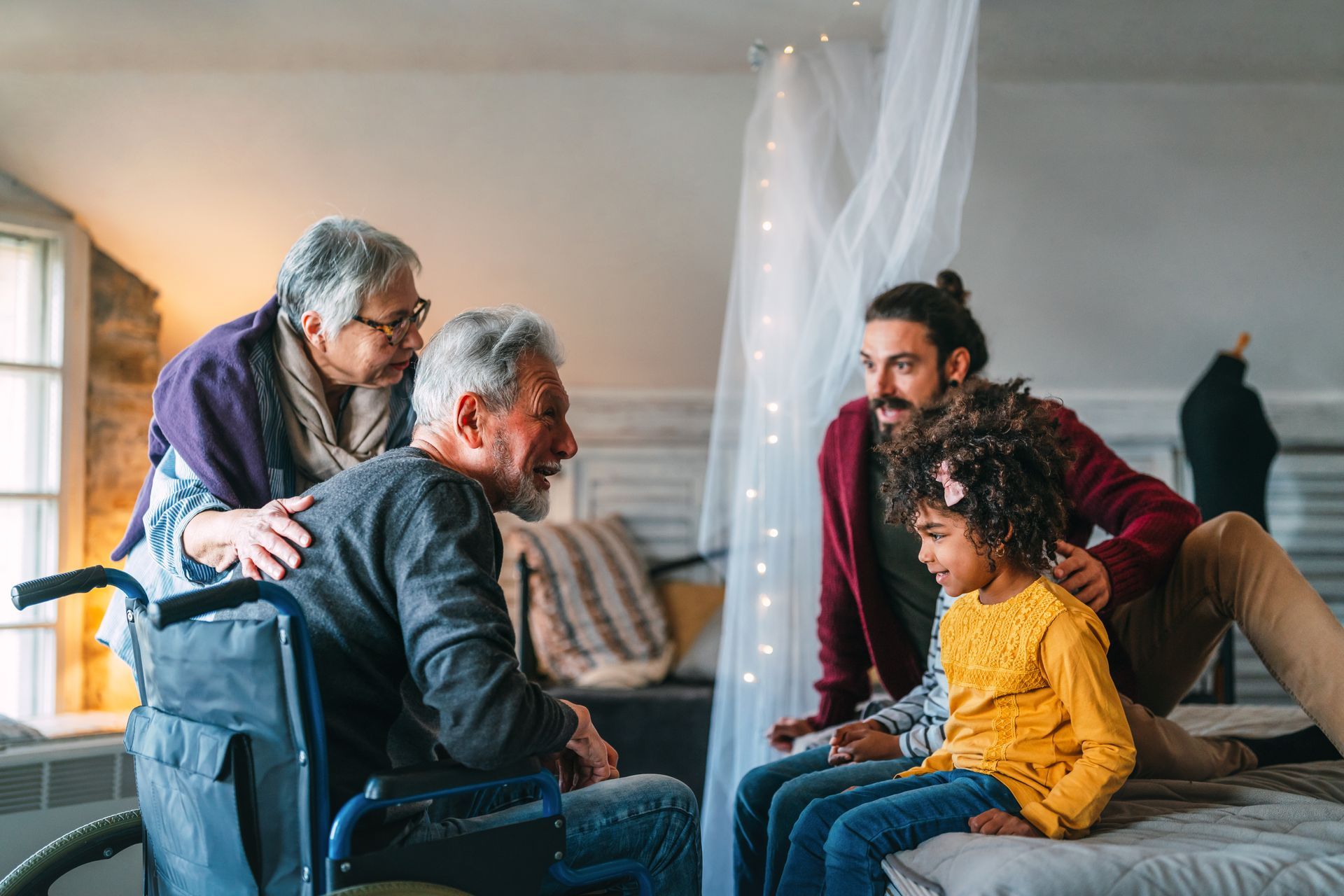 Family gathering around a man in a wheelchair; a woman has her hands on his shoulders; a man and child sit on a bed.