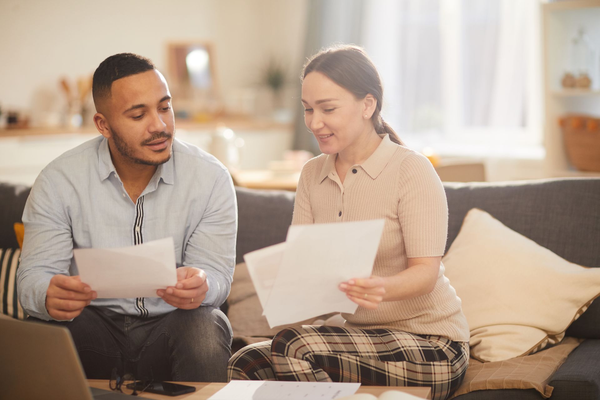 Couple reviewing documents on a couch indoors. Man looks thoughtful, woman smiles. Sunlight streams in.