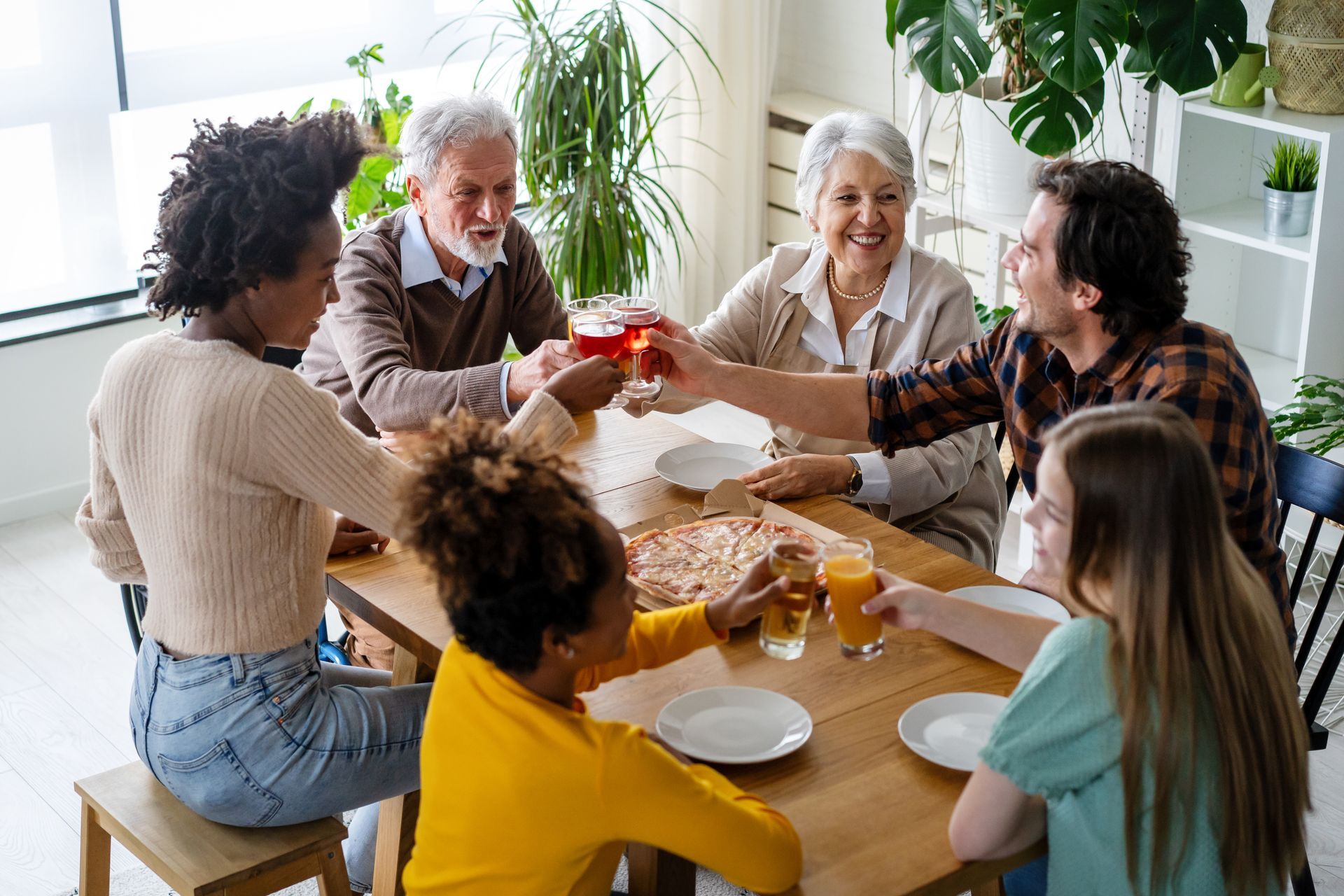 Family toasting drinks around a dining table with pizza and plants.