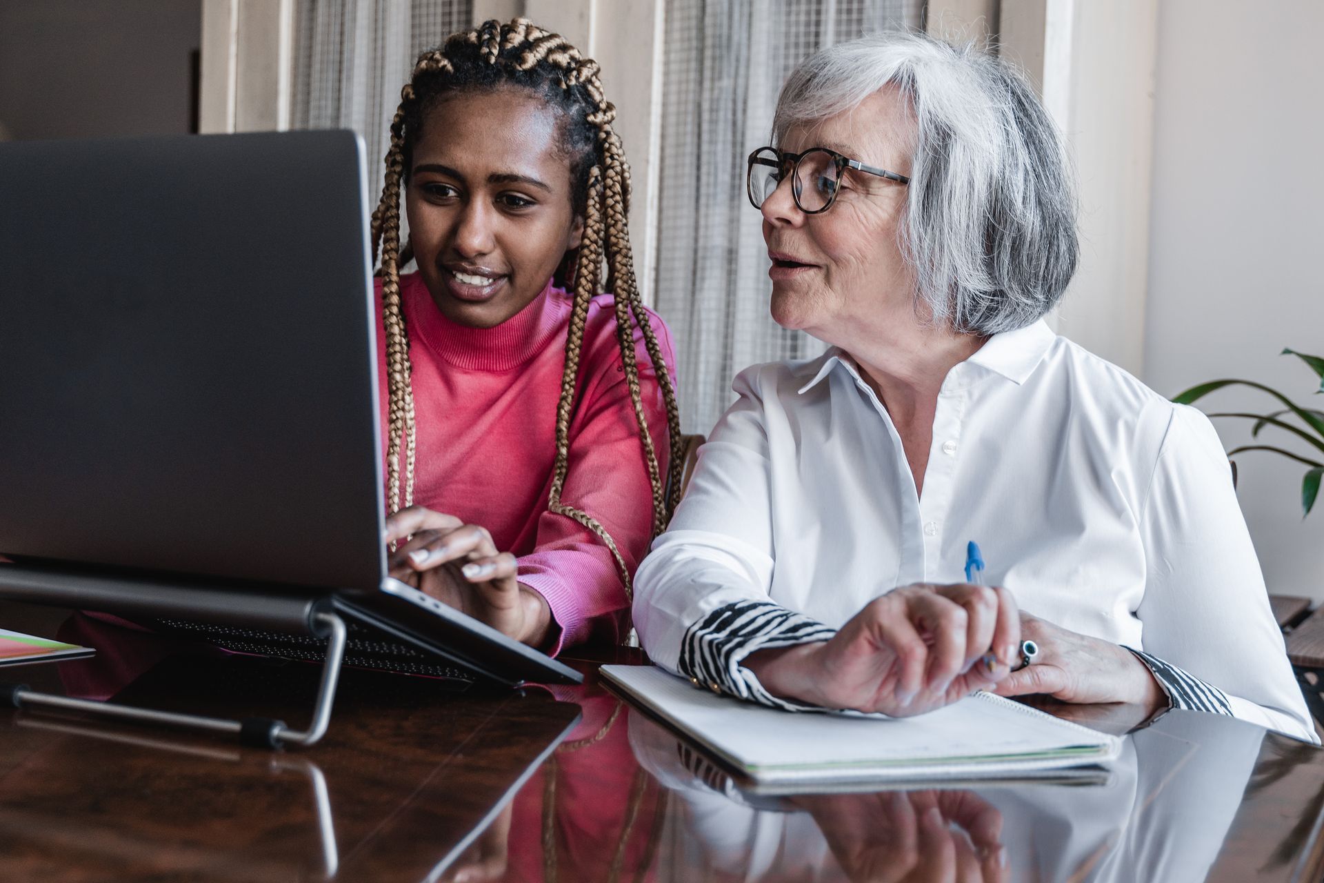 Woman with braids shows laptop to another woman at a table, both looking at the screen.