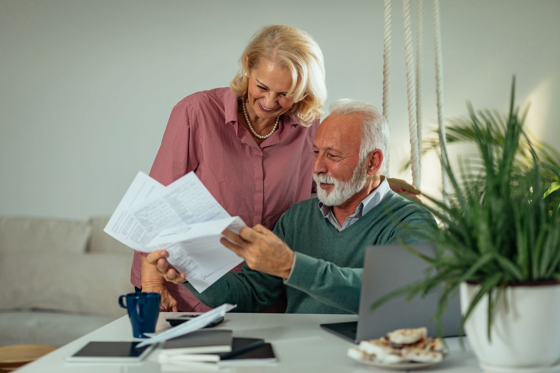 Senior couple reviewing documents together at a table with a laptop, smiles, and indoor plants.
