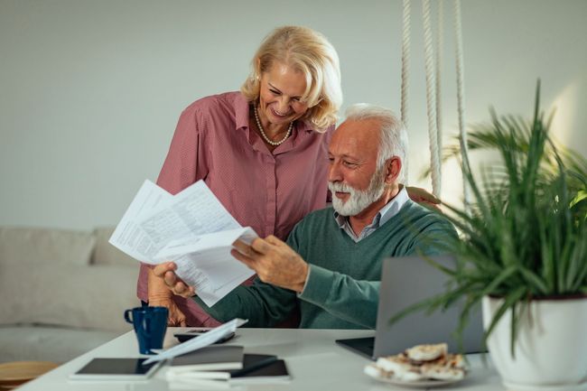 Senior couple reviewing documents together at a table with a laptop, smiles, and indoor plants.
