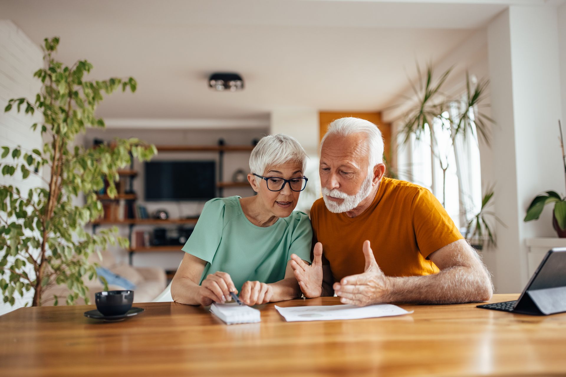 An older couple reviews documents and uses a calculator at a wooden table in a well-lit living room.