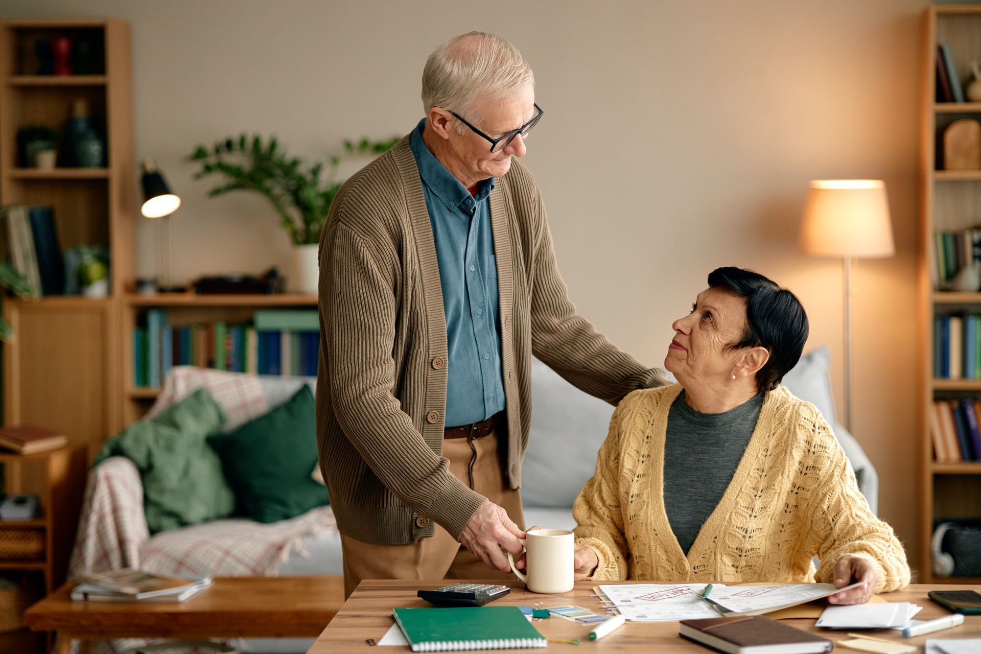 Older man offers coffee to a woman working at a table, home interior.