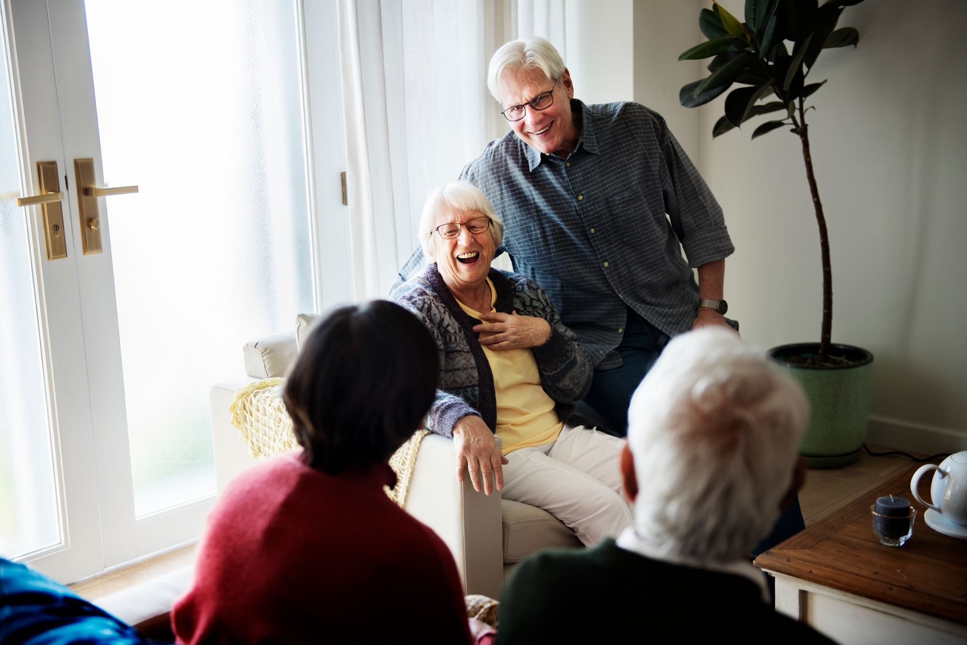 Family exchanging a gift. A girl gives flowers to a woman, seated. Others watch, smiling, by a window.