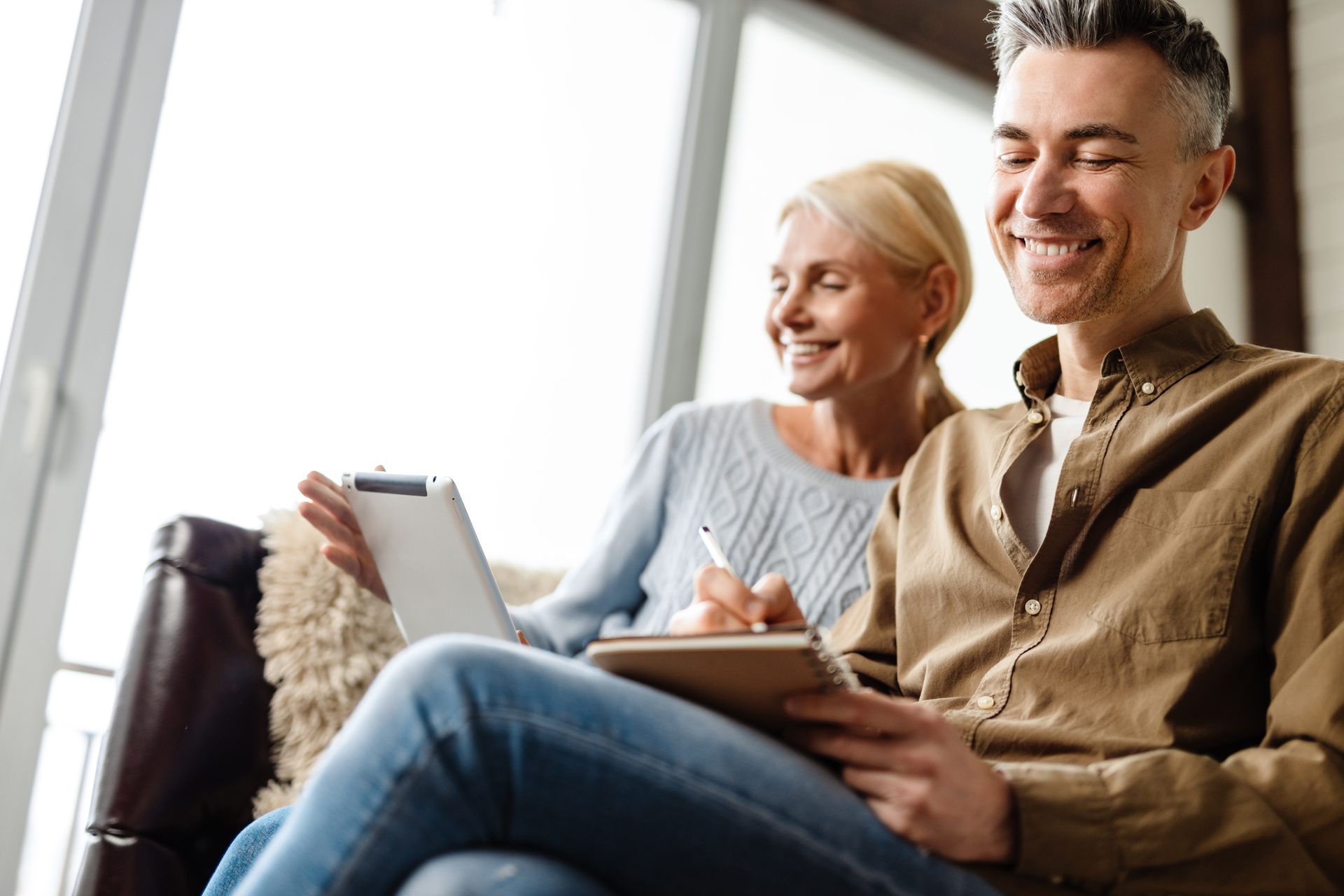 Man smiling, writing on a notepad, with woman looking at a tablet in front of a window.