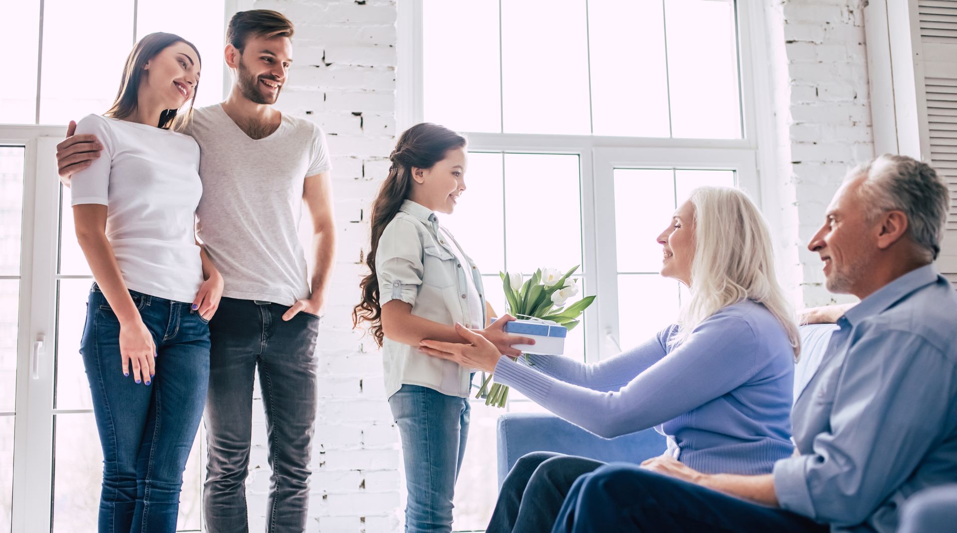 Family exchanging a gift. A girl gives flowers to a woman, seated. Others watch, smiling, by a window.