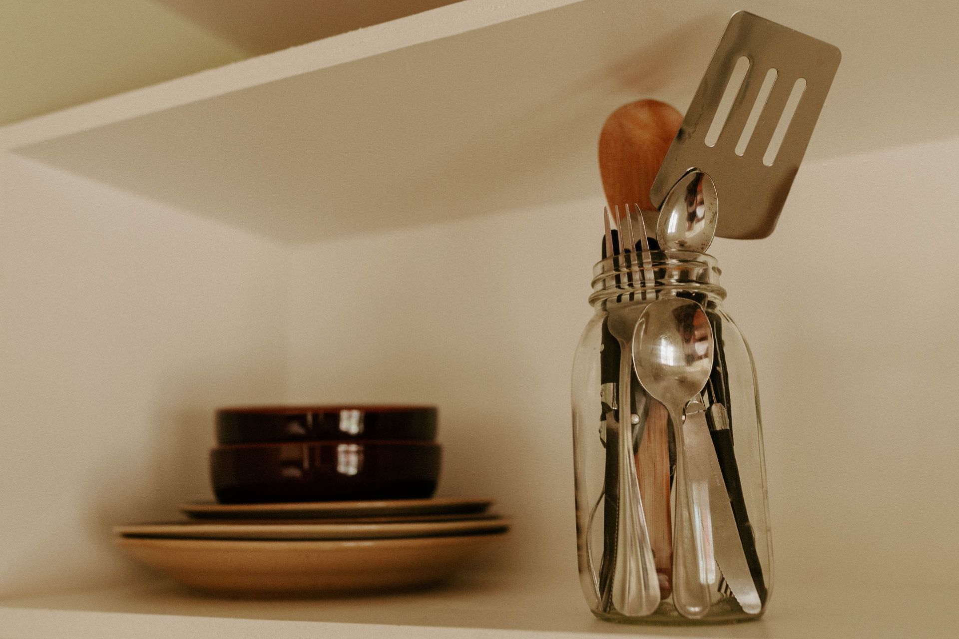 A jar filled with silverware and a spatula on a shelf.