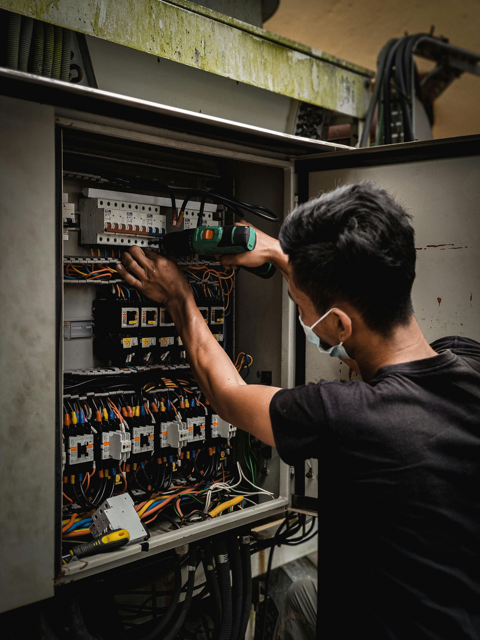 A person wearing a mask works on an electrical panel with a power drill in an industrial setting.