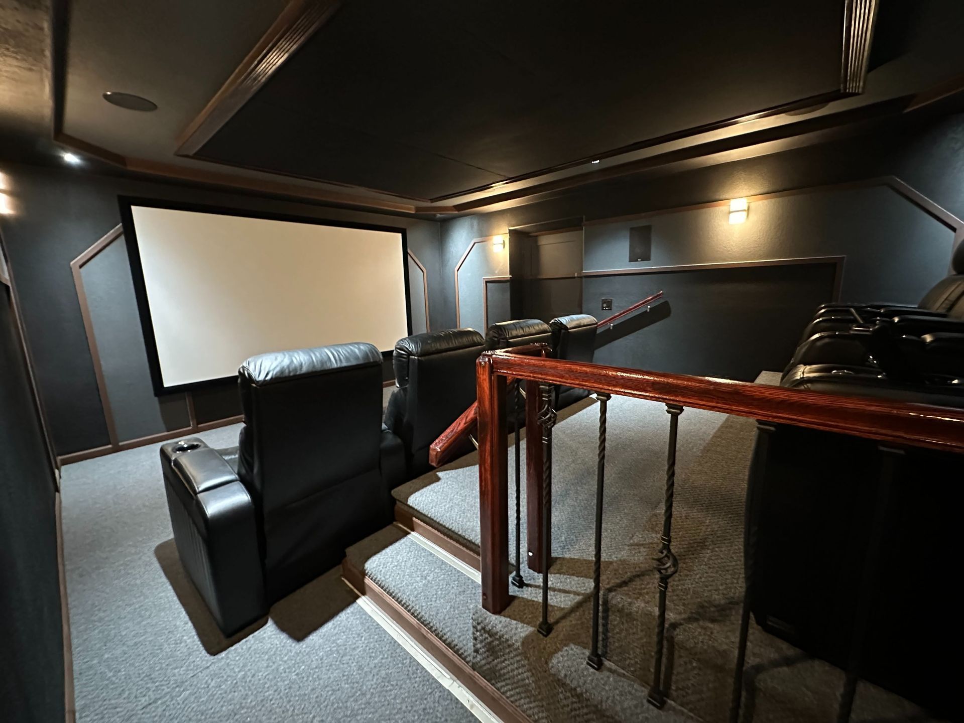 Small home theater with black recliners, large screen, and staircase railing in dim lighting
