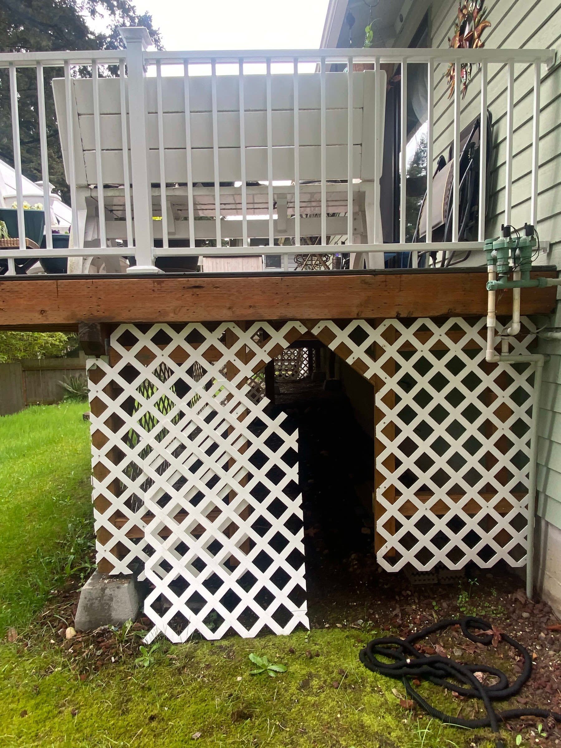 Under-deck storage area with white lattice, black trash bin, and coiled hose on grass