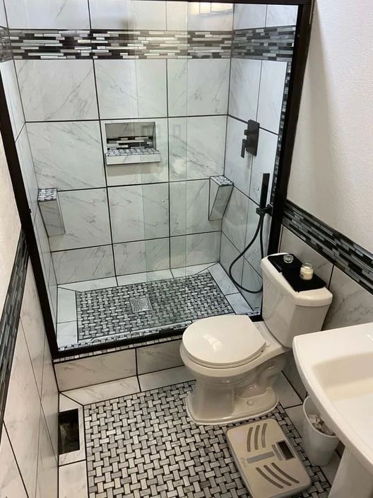 Small tiled bathroom with glass shower, toilet, sink, and patterned shower floor mat.