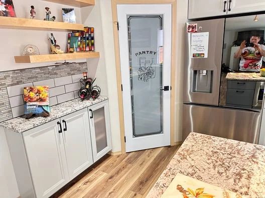 Bright kitchen with white cabinets, stainless fridge, open shelves, and a frosted glass pantry door