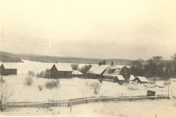 A black and white photo of a snowy village with a fence in the foreground.