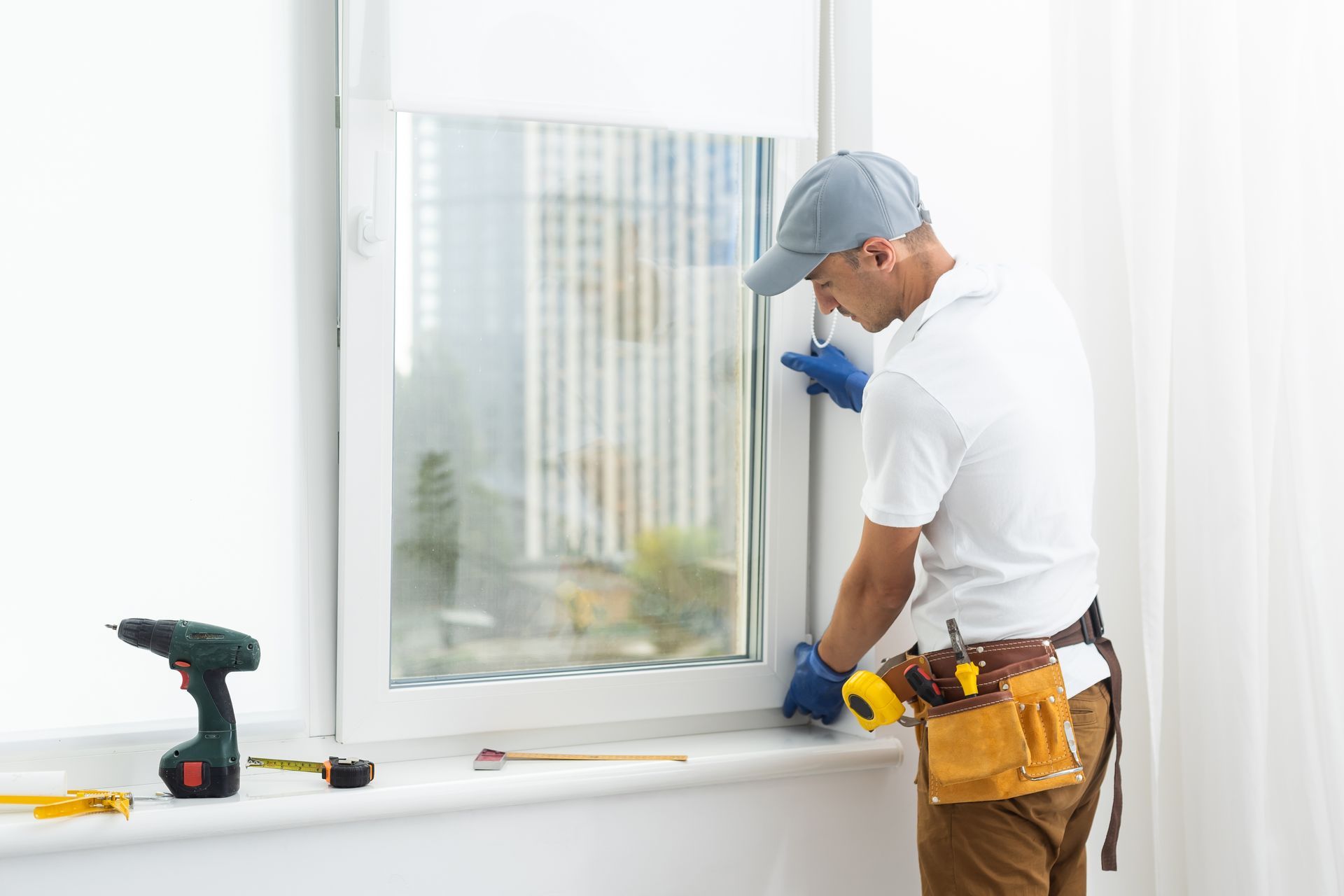 A man installing a window, wearing a cap, gloves, and tool belt, indoors with a bright window.