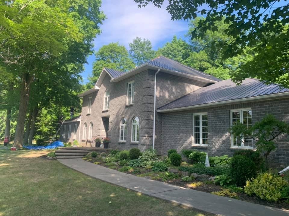 Gray brick house with white-framed windows and a dark roof, set among trees and a green lawn.