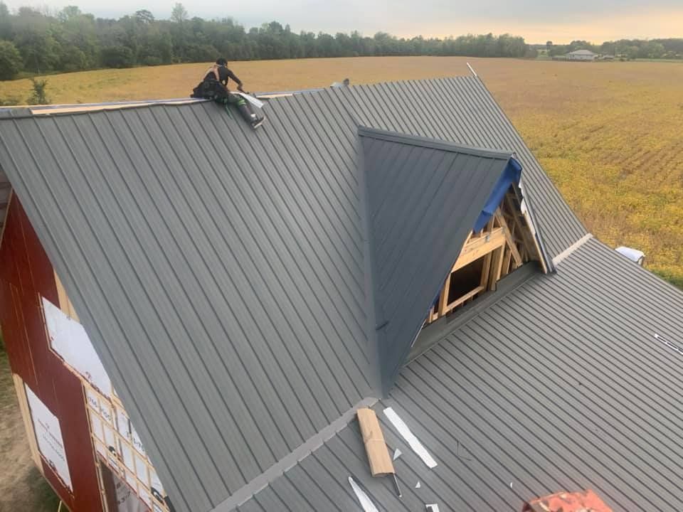 Person on a gray metal roof installing panels on a red barn with a blue gable, rural setting.