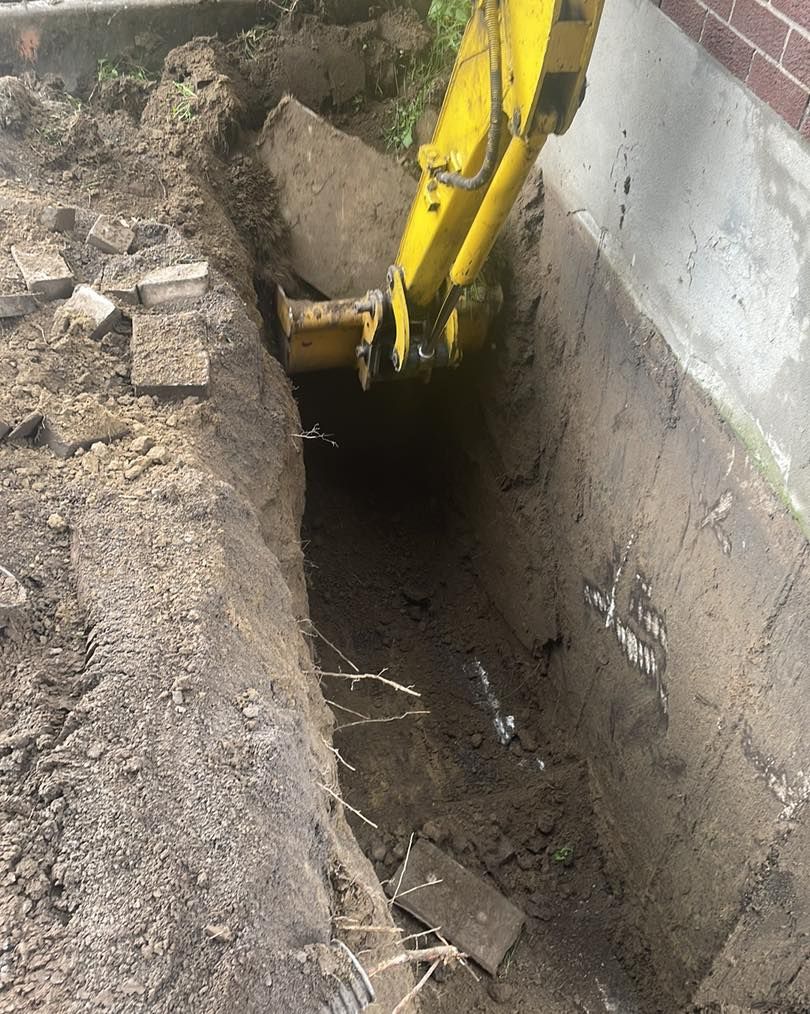 A yellow excavator digging a trench beside a concrete building foundation.