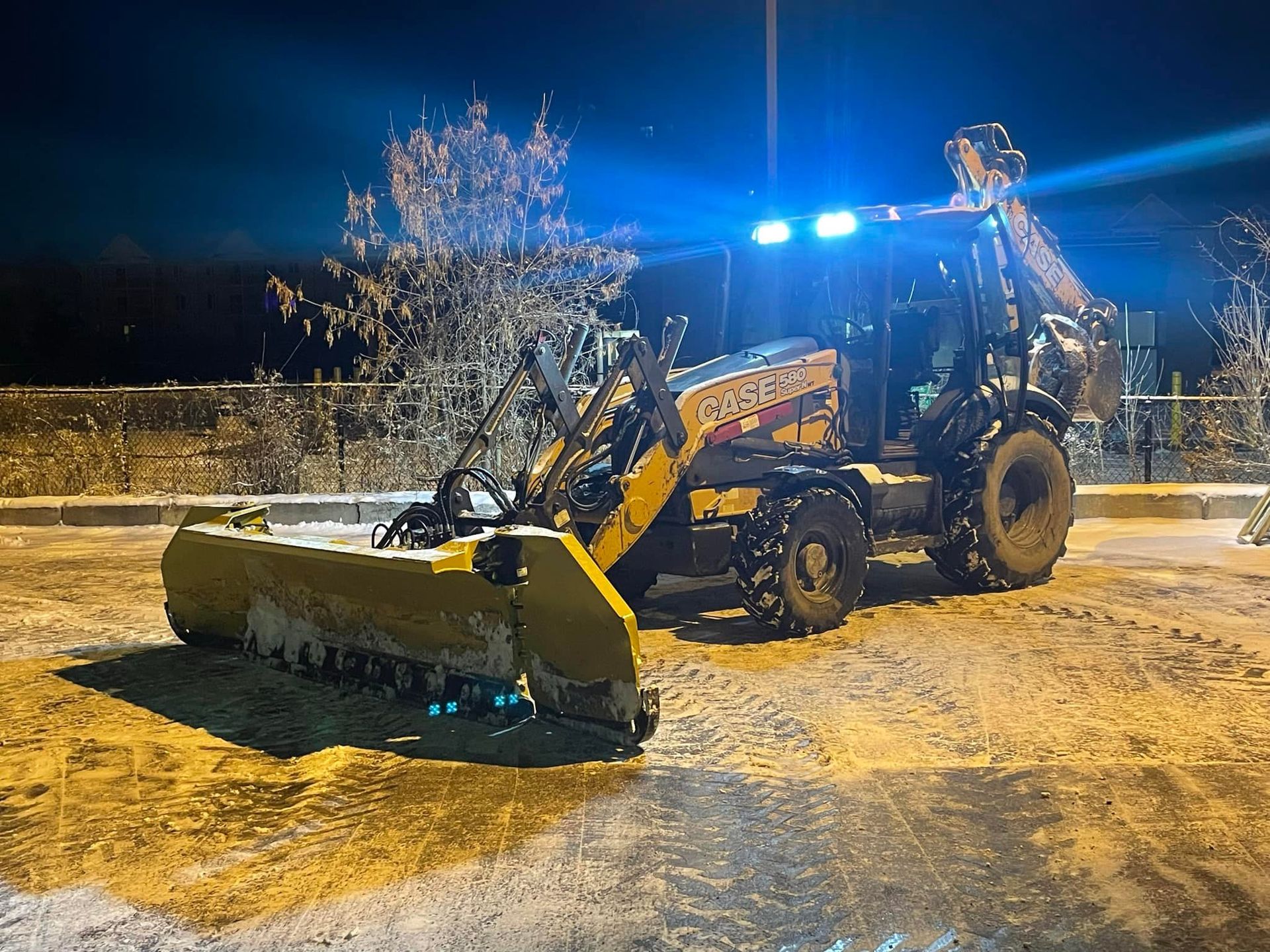 Yellow snowplow clearing a snowy area at night, with bright blue lights.