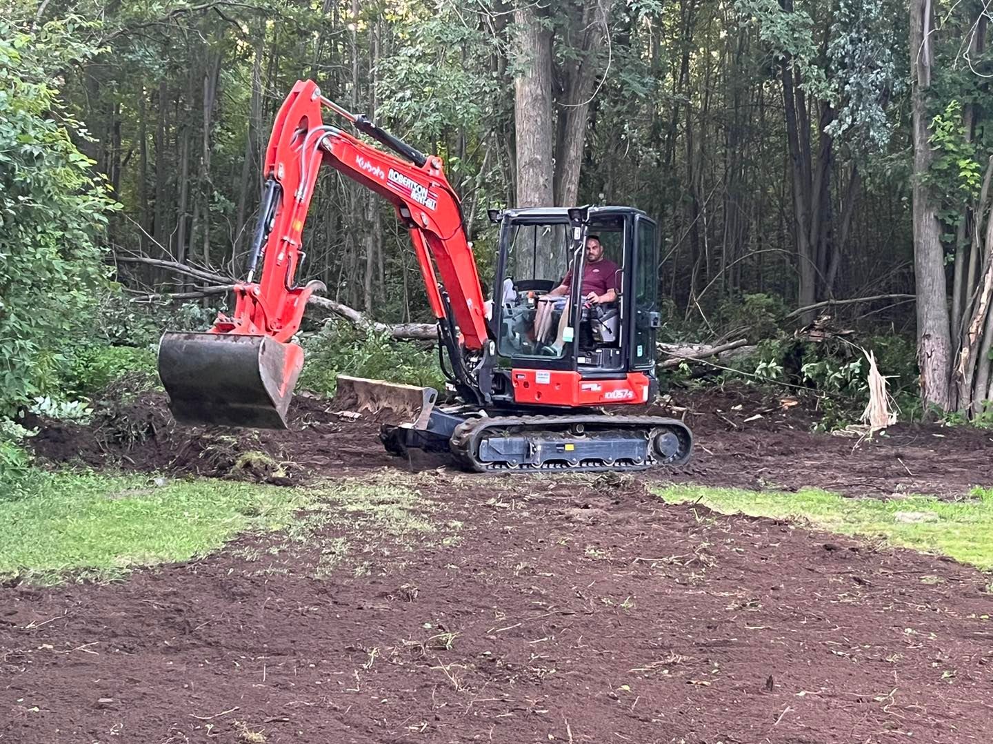 Red excavator digging in a dirt clearing, operator inside, surrounded by trees.