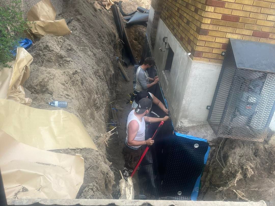 Two workers installing a black waterproof membrane along a brick foundation wall in an excavated trench.
