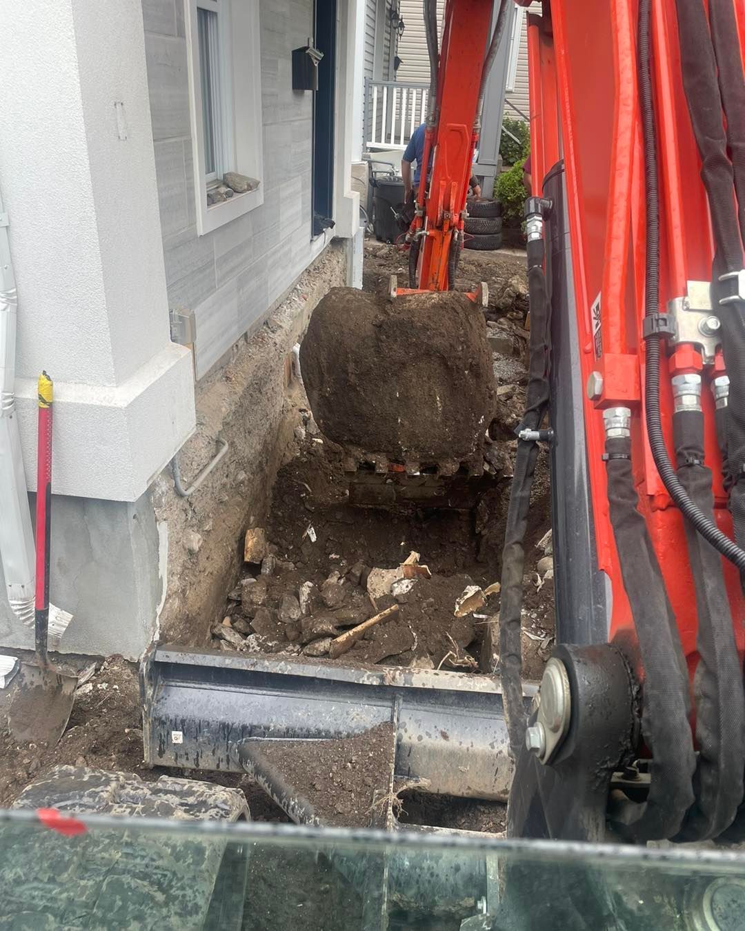 Excavator digging trench next to a building's foundation; orange machinery, gray siding.