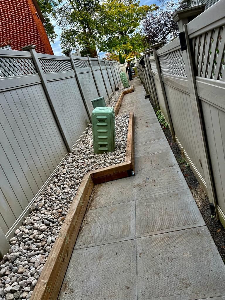 Narrow walkway between fences, with gravel bed and utility boxes.