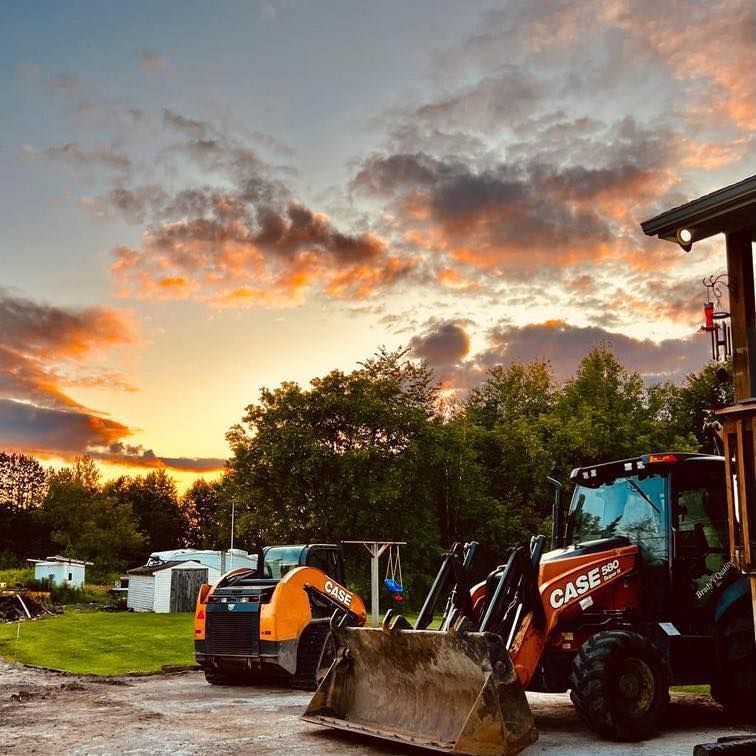 Construction equipment parked outdoors at sunset; orange and red sky, trees and small buildings in the background.