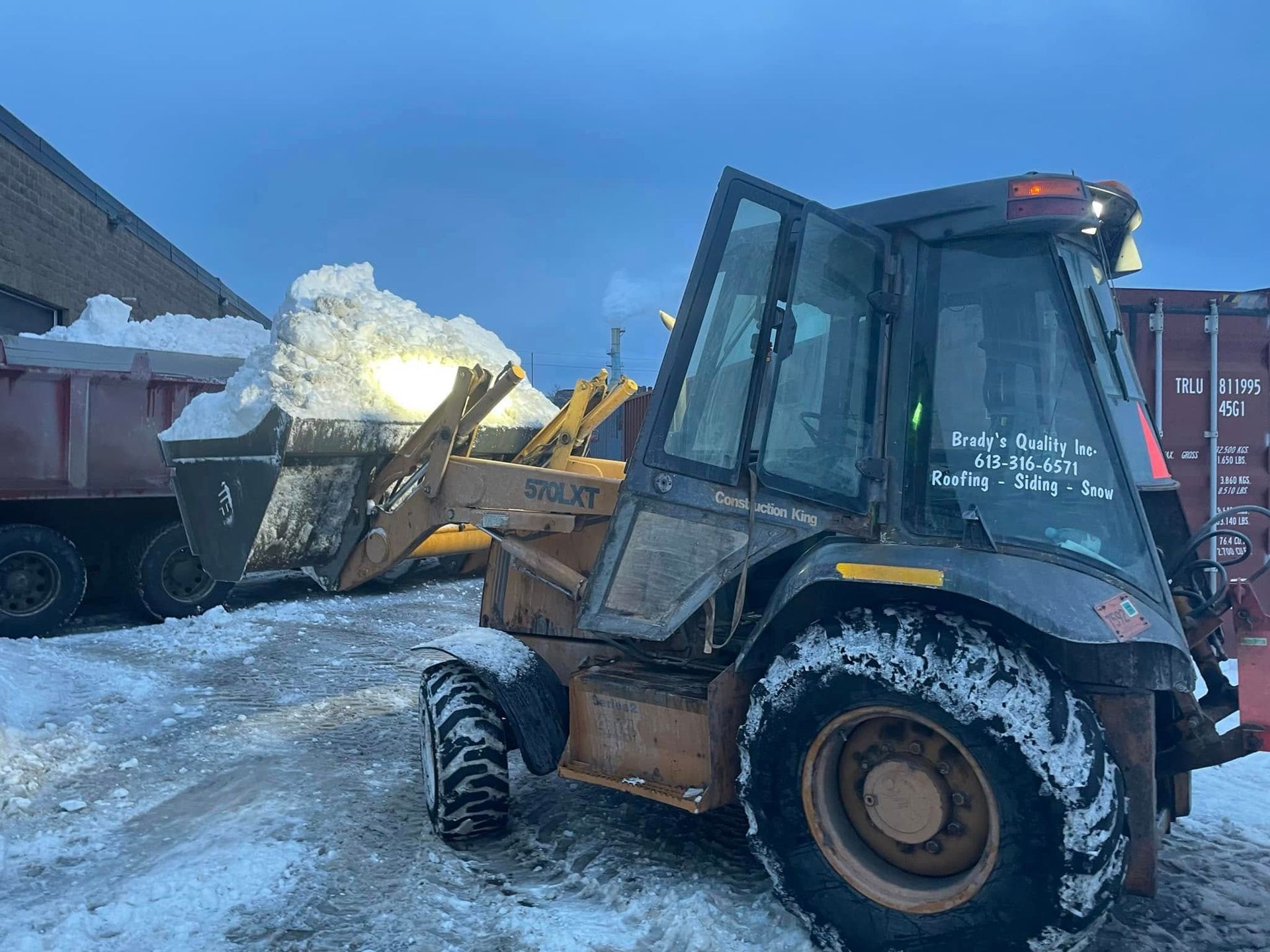 A backhoe loader scoops snow into a dump truck in a snowy outdoor setting.