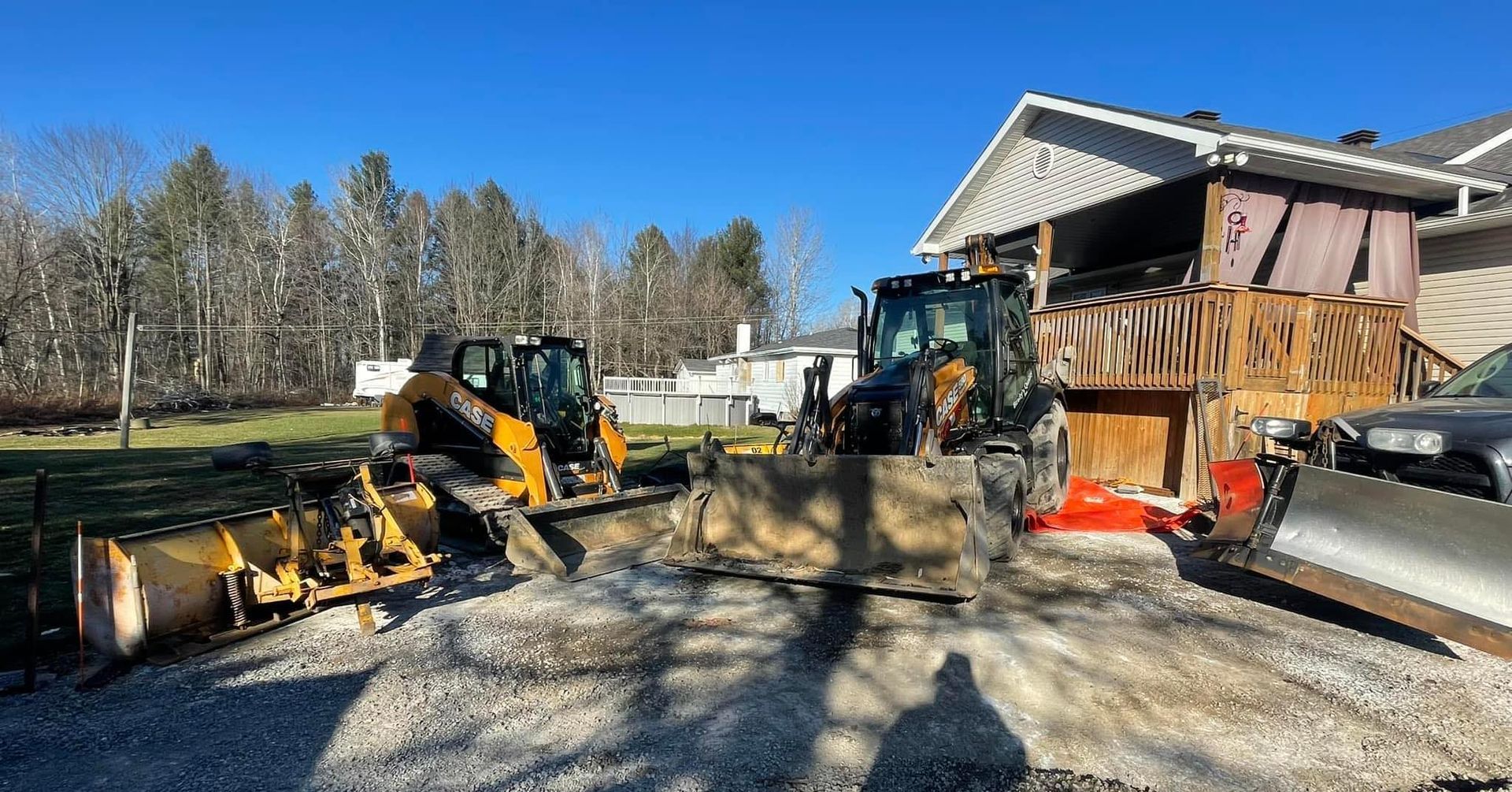 Two construction vehicles clearing gravel driveway next to house under blue sky.