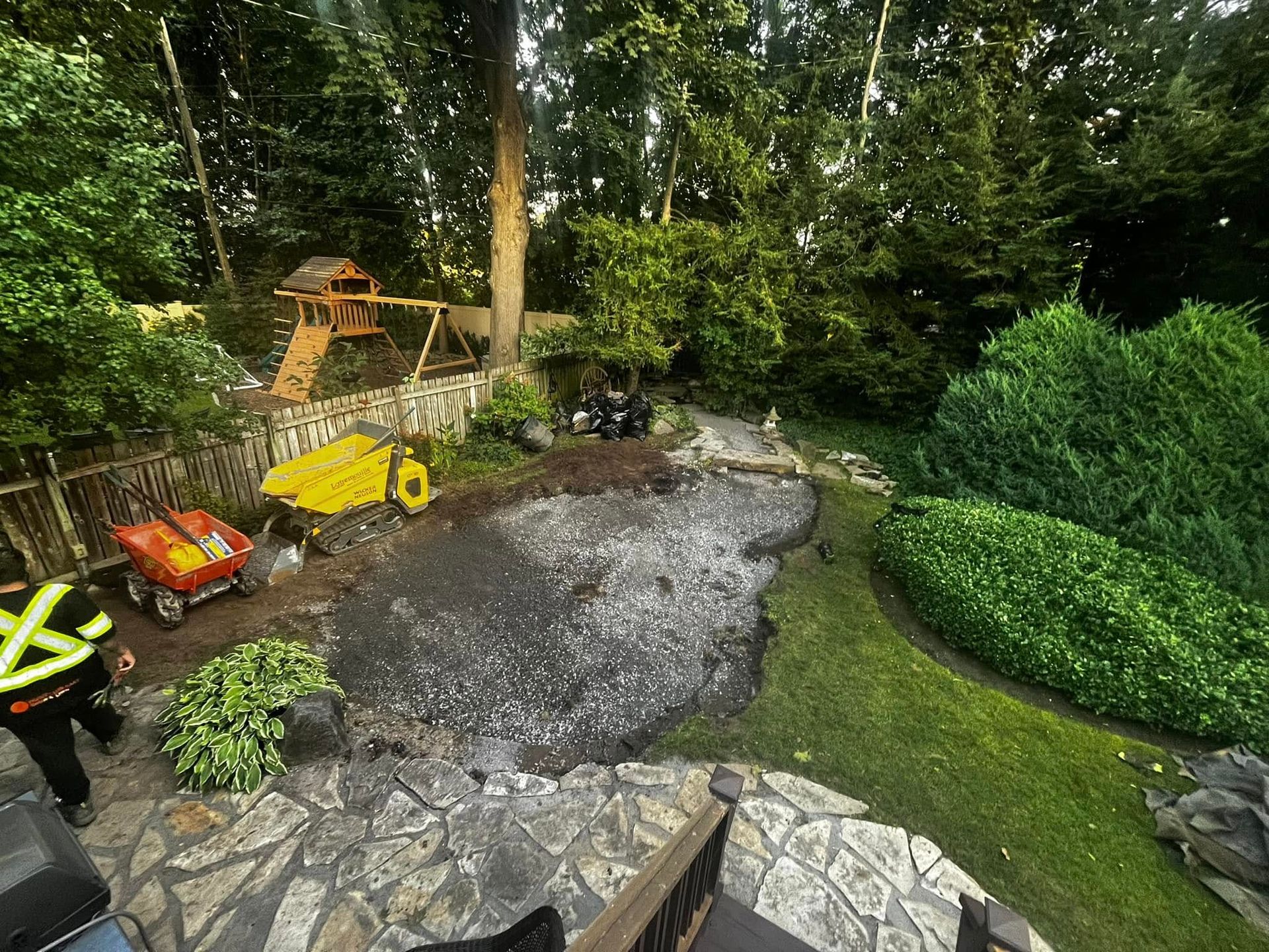Backyard with pond construction. Gravel-filled space, machinery, worker in safety vest, playground in background.