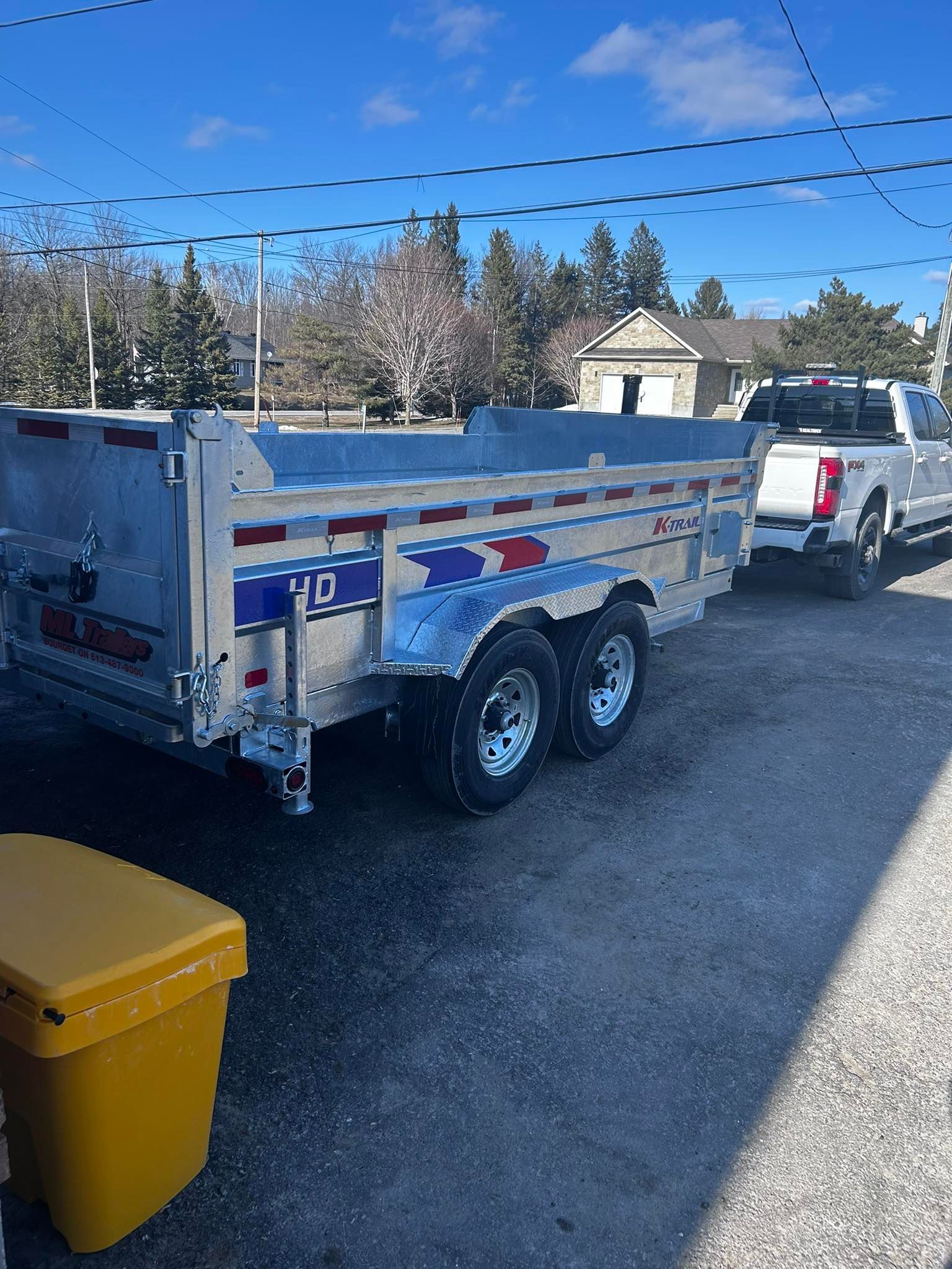 A silver dump trailer hitched to a white pickup truck, parked on pavement; a yellow bin is in front.