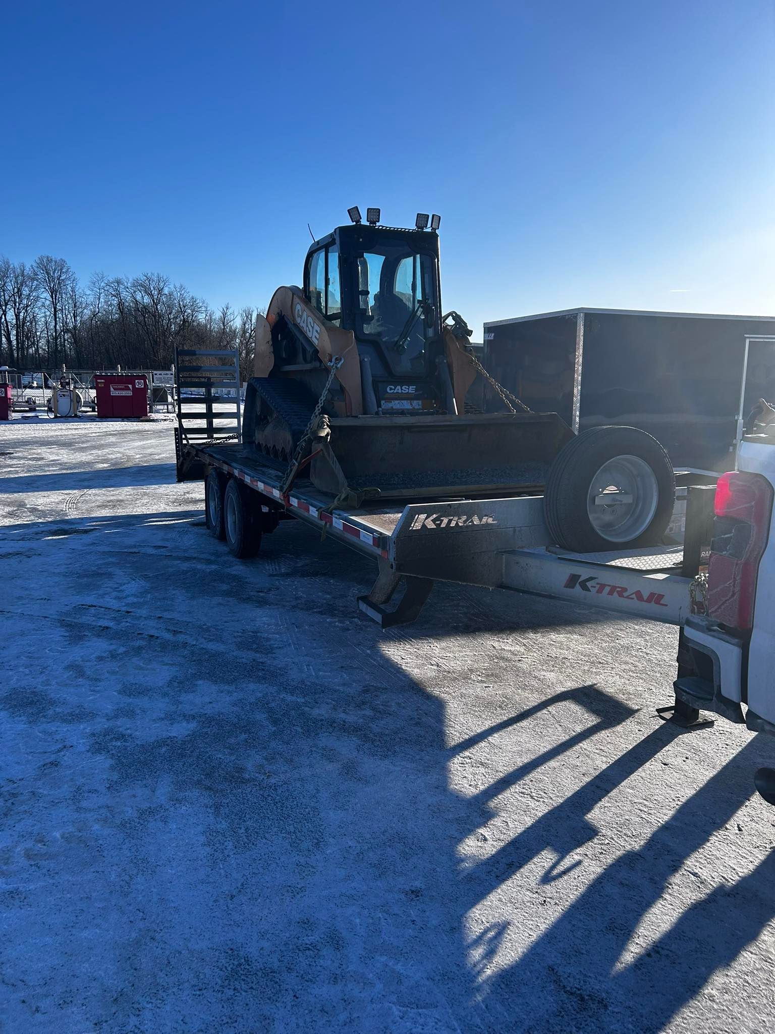 Backhoe on a trailer in a snowy outdoor setting under a clear, sunny sky.