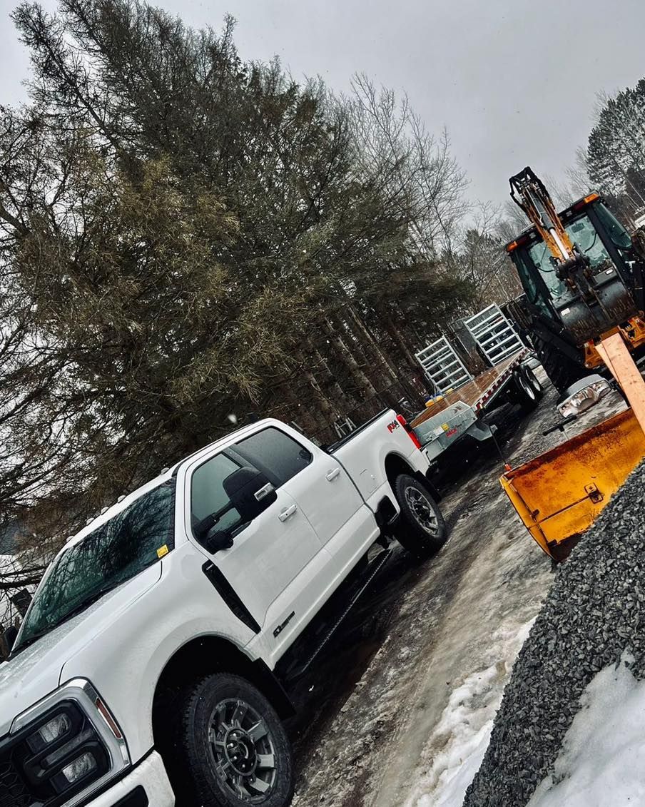 White truck and tractor parked near snow-covered gravel, next to some trees, with a cloudy sky.