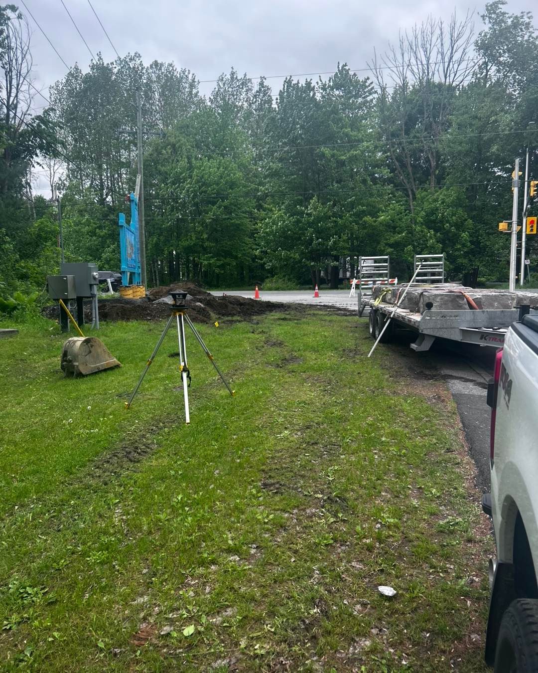 Construction site. A truck, a trailer, and a digging machine sit on a grassy area near a road.