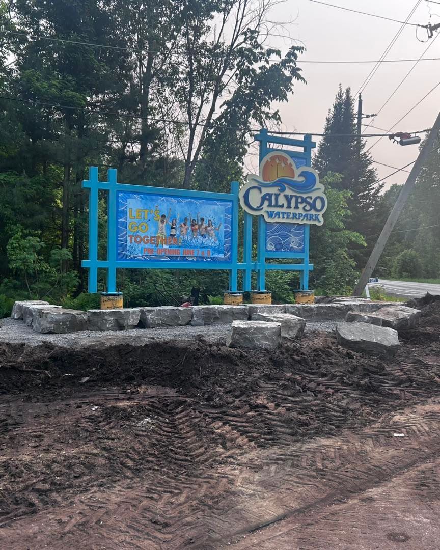 Calypso Waterpark sign, blue and white, on a stone base, with dirt in front; trees and sky in the background.