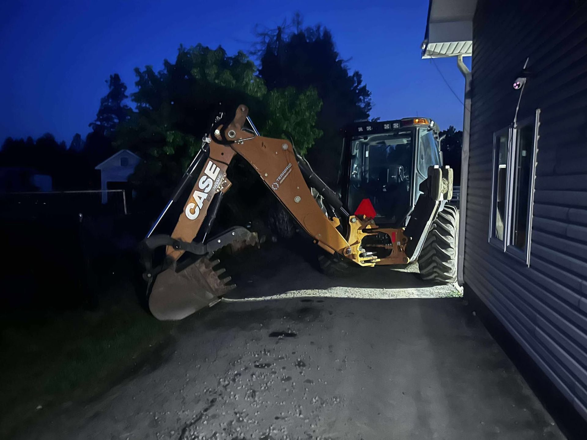 A Case backhoe parked on a gravel driveway next to a house at dusk.