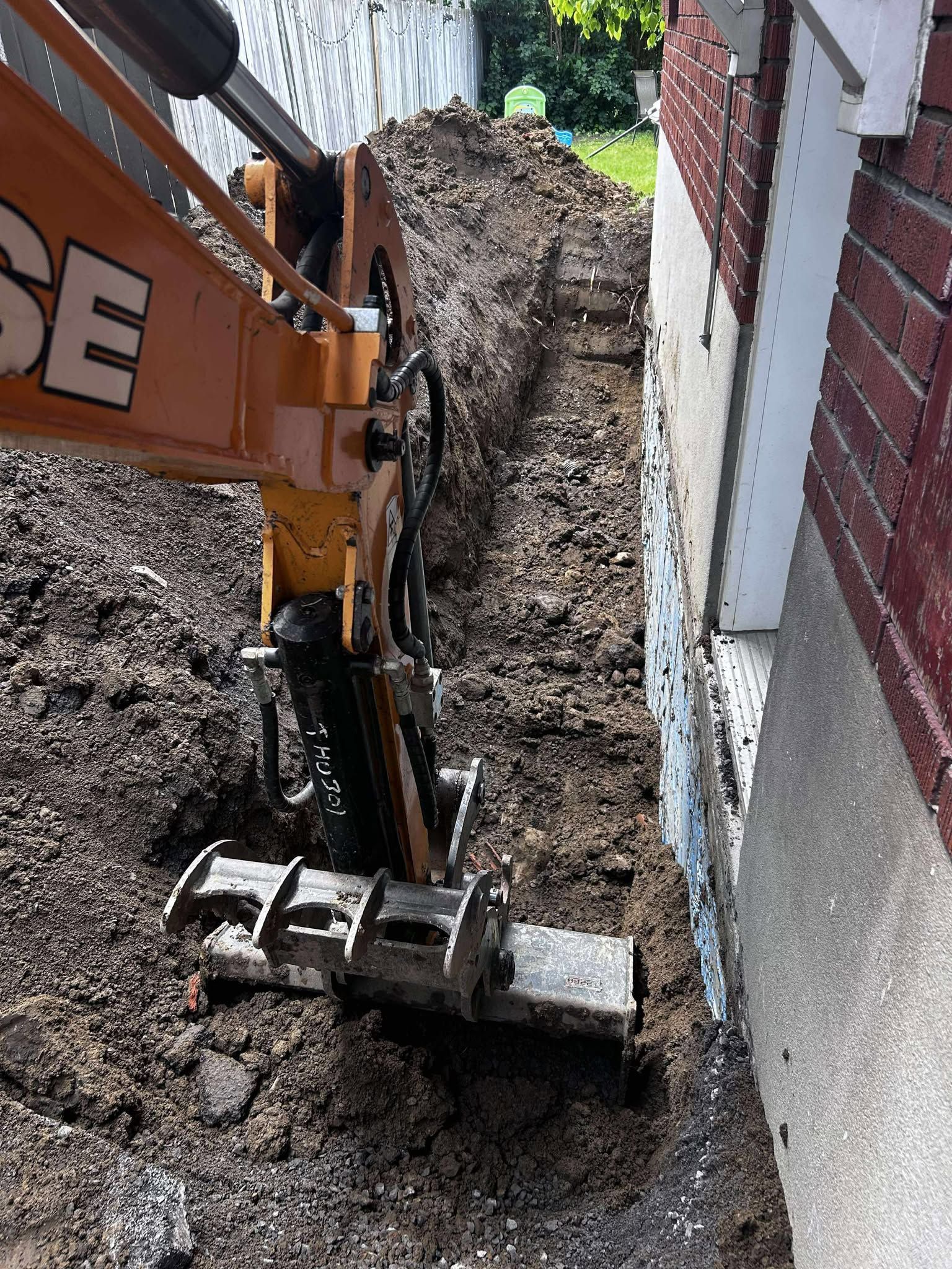 An orange excavator digging a trench next to a brick building. Earth is piled up.