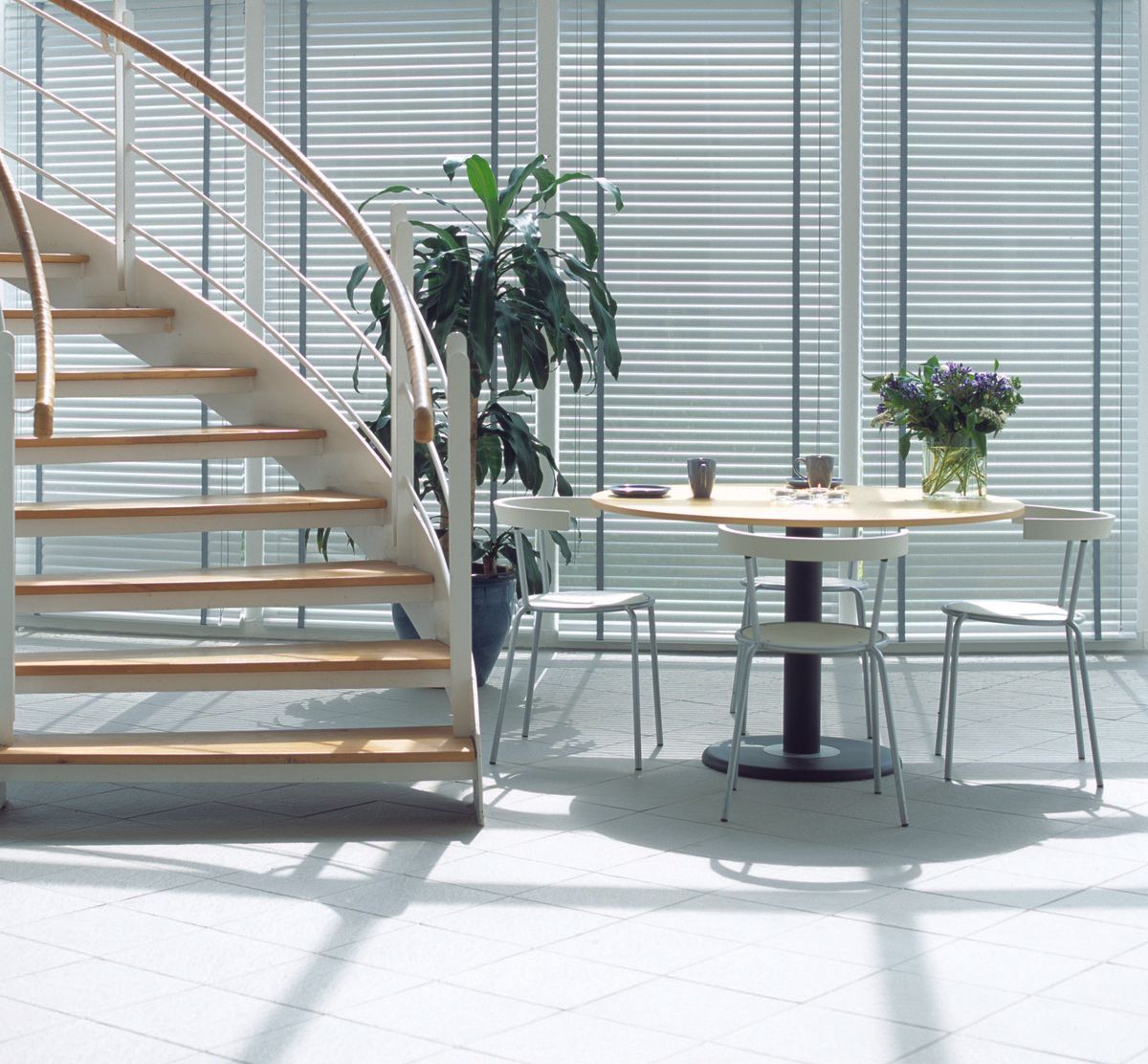 White spiral staircase, table with chairs, large windows with blinds, and potted plant in a bright room.
