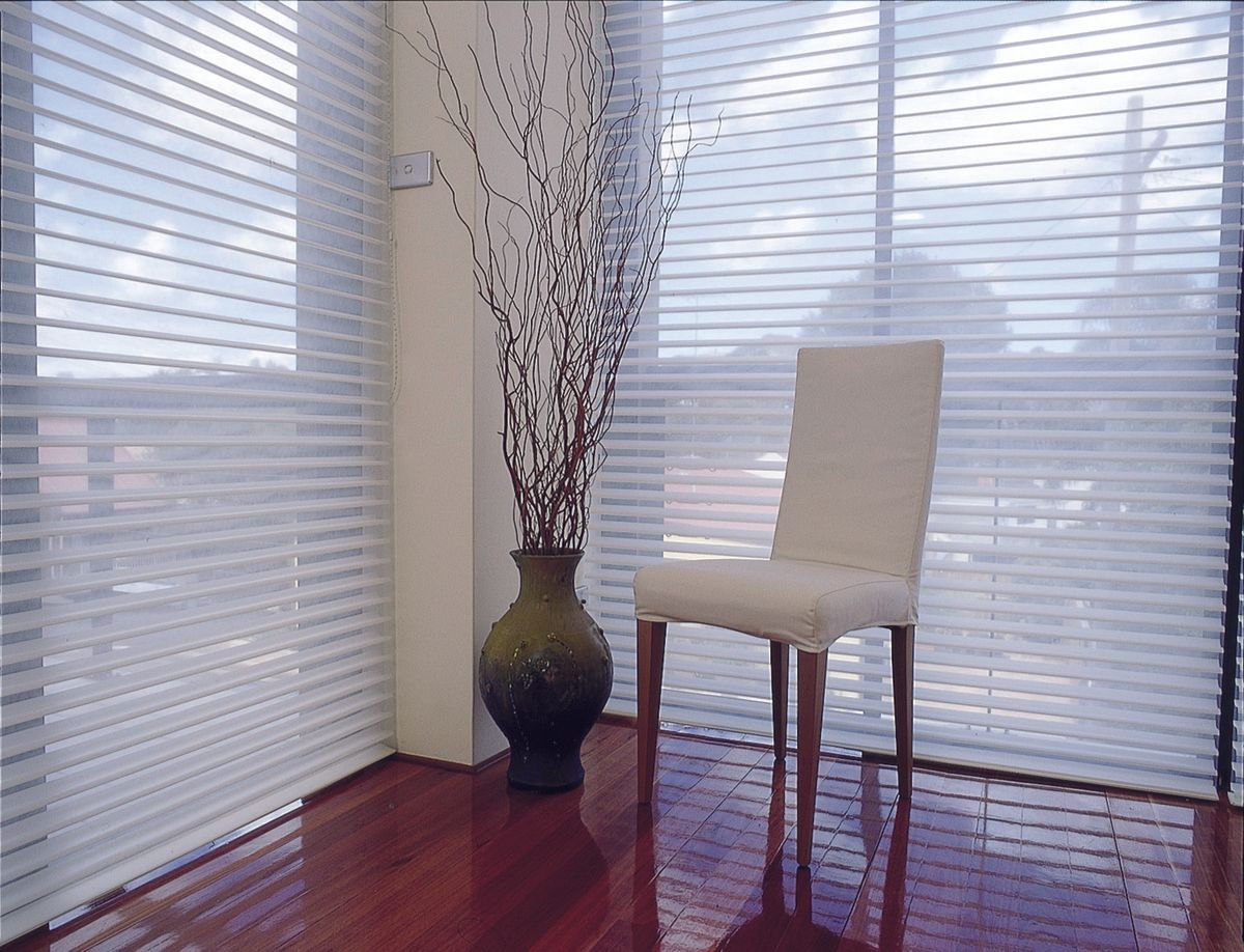 Corner room with sheer white blinds, dark wood floors, a chair, and a vase with branches.