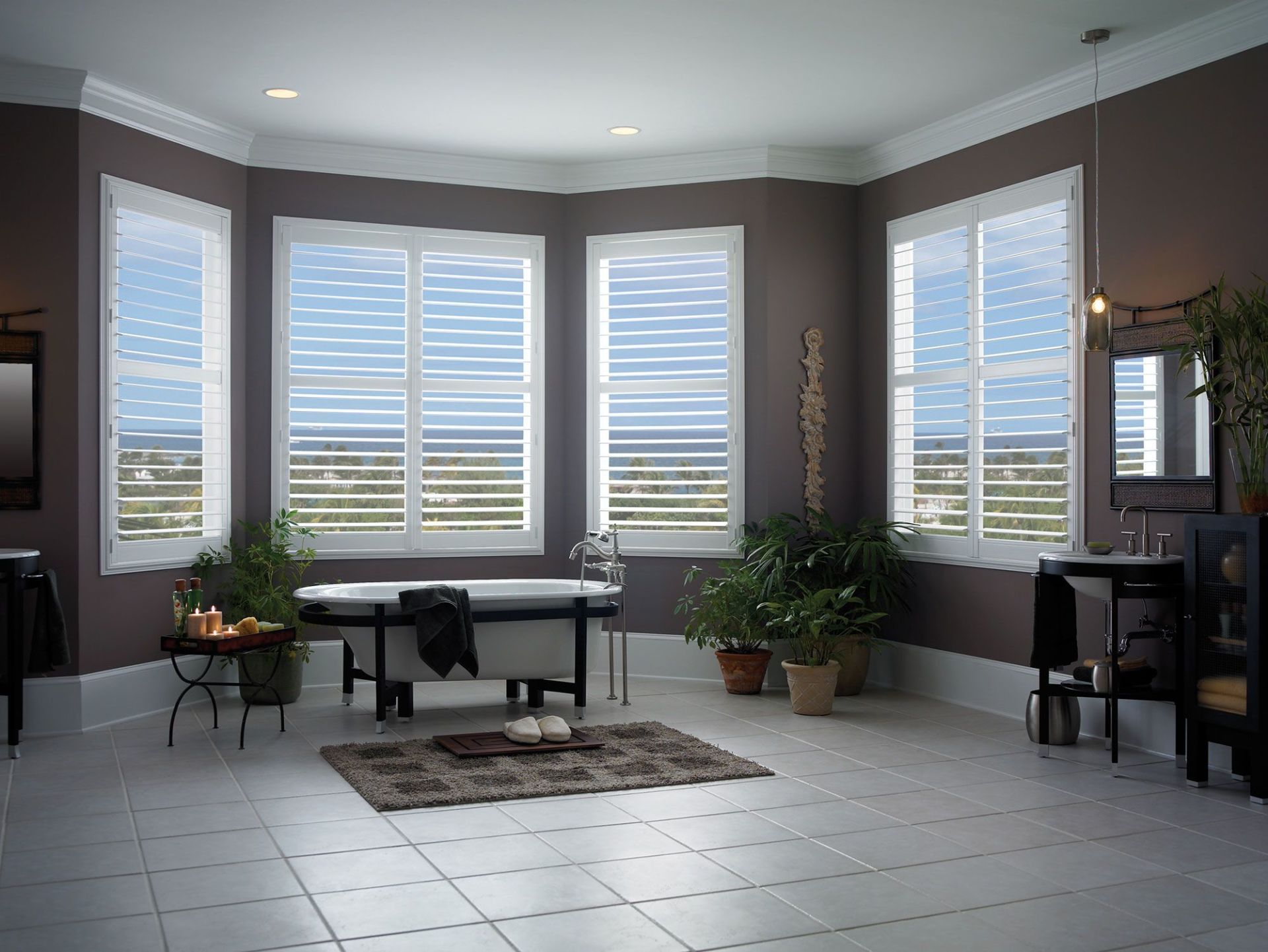 Bathroom with white tile floor, bathtub under large windows with shutters, and potted plants.