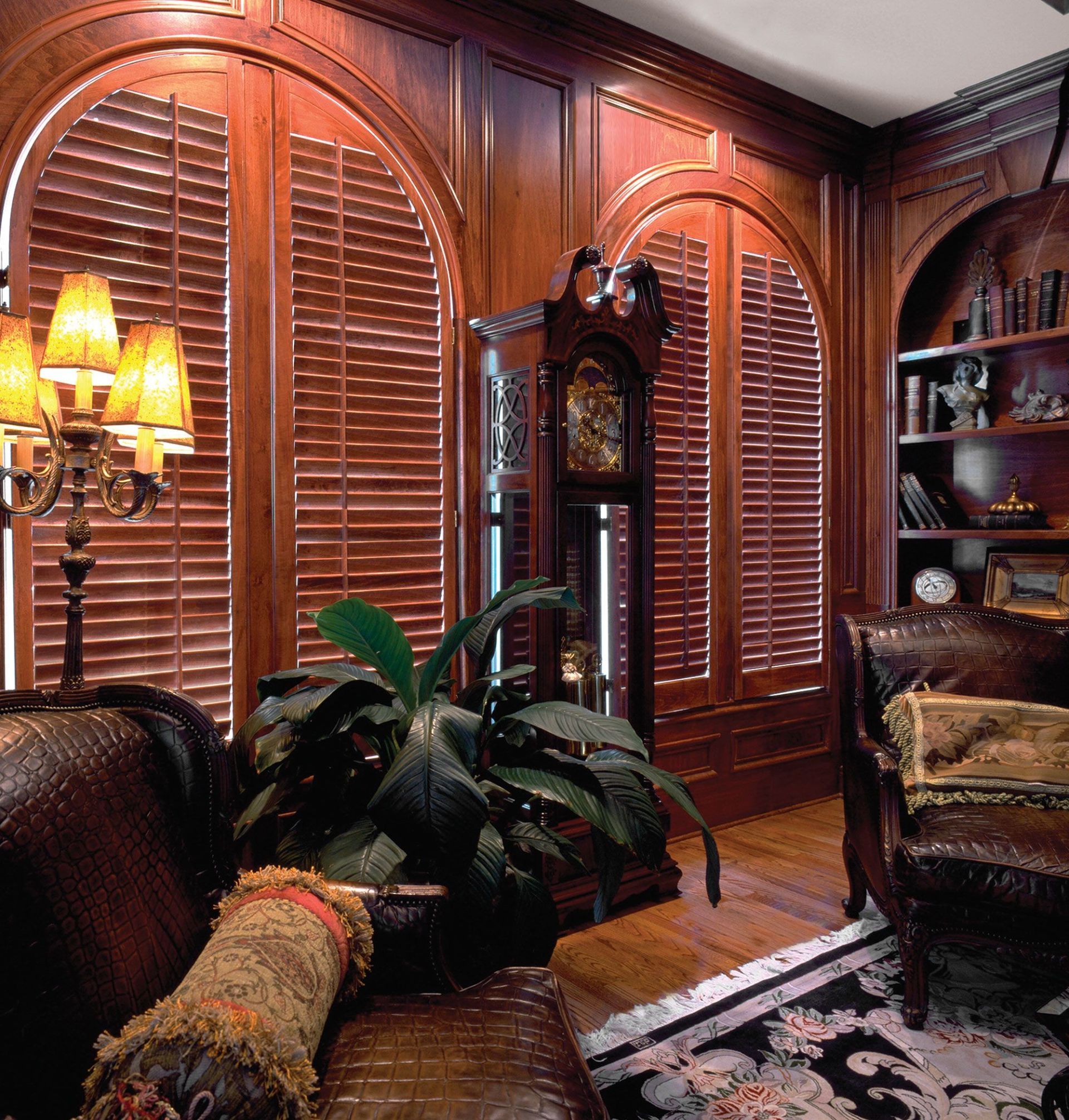 Elegant wood-paneled room with arched shuttered windows, grandfather clock, leather furniture, and a large plant.