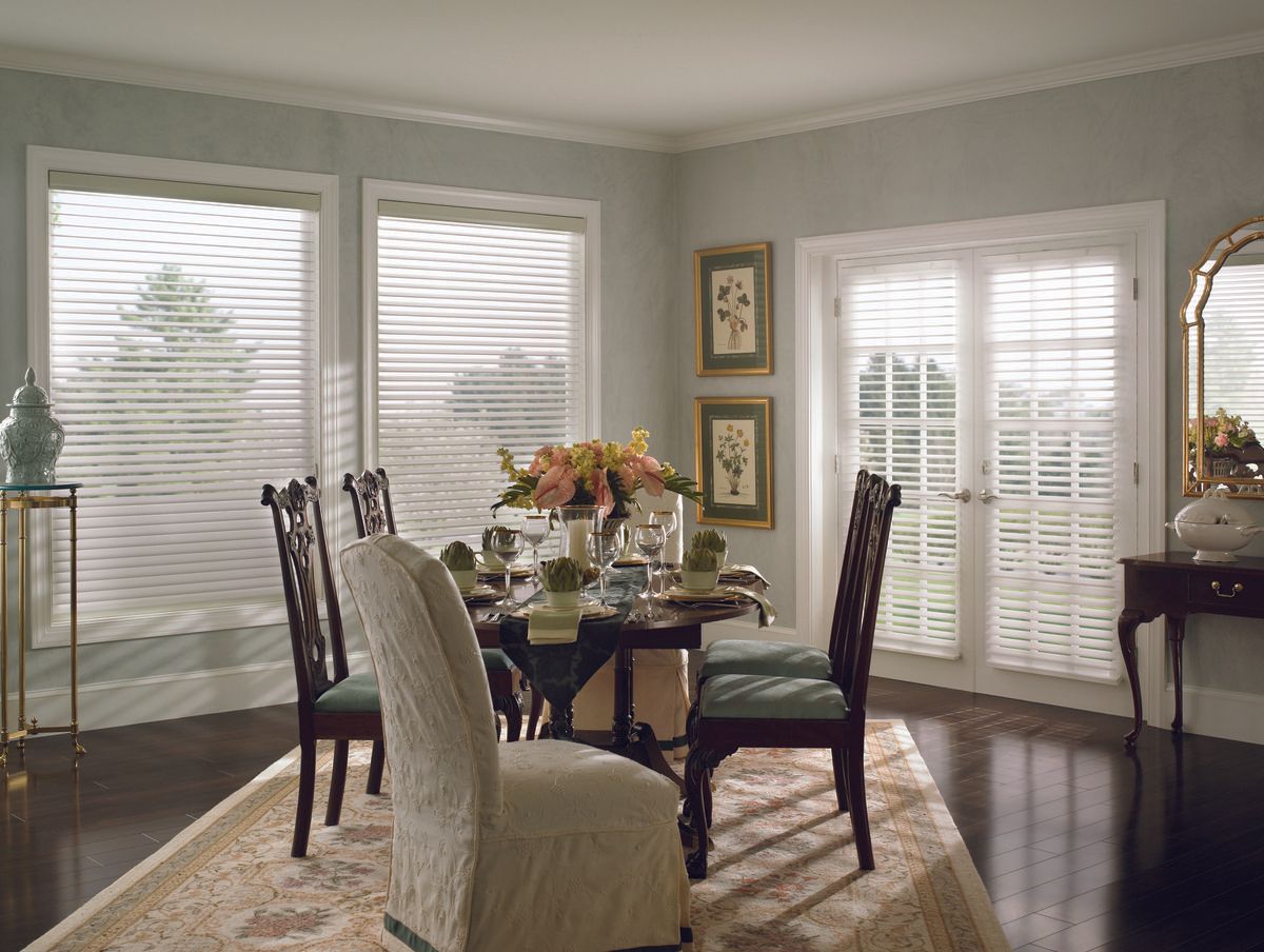 Dining room with round table, chairs, and blinds on windows. Green walls, dark wood floor, and floral centerpiece.