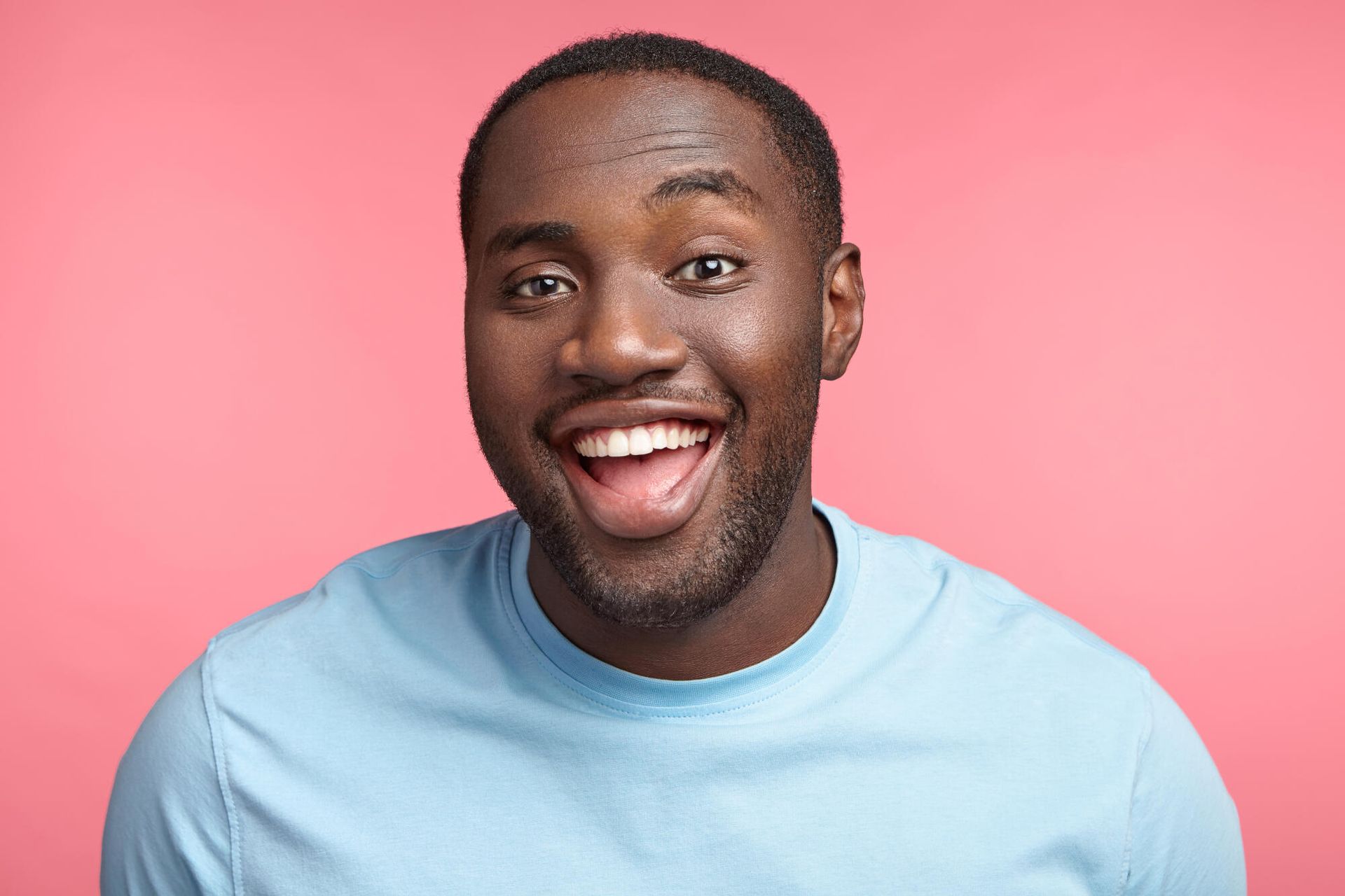 Smiling person with dark skin against a pink background, wearing a light blue shirt.