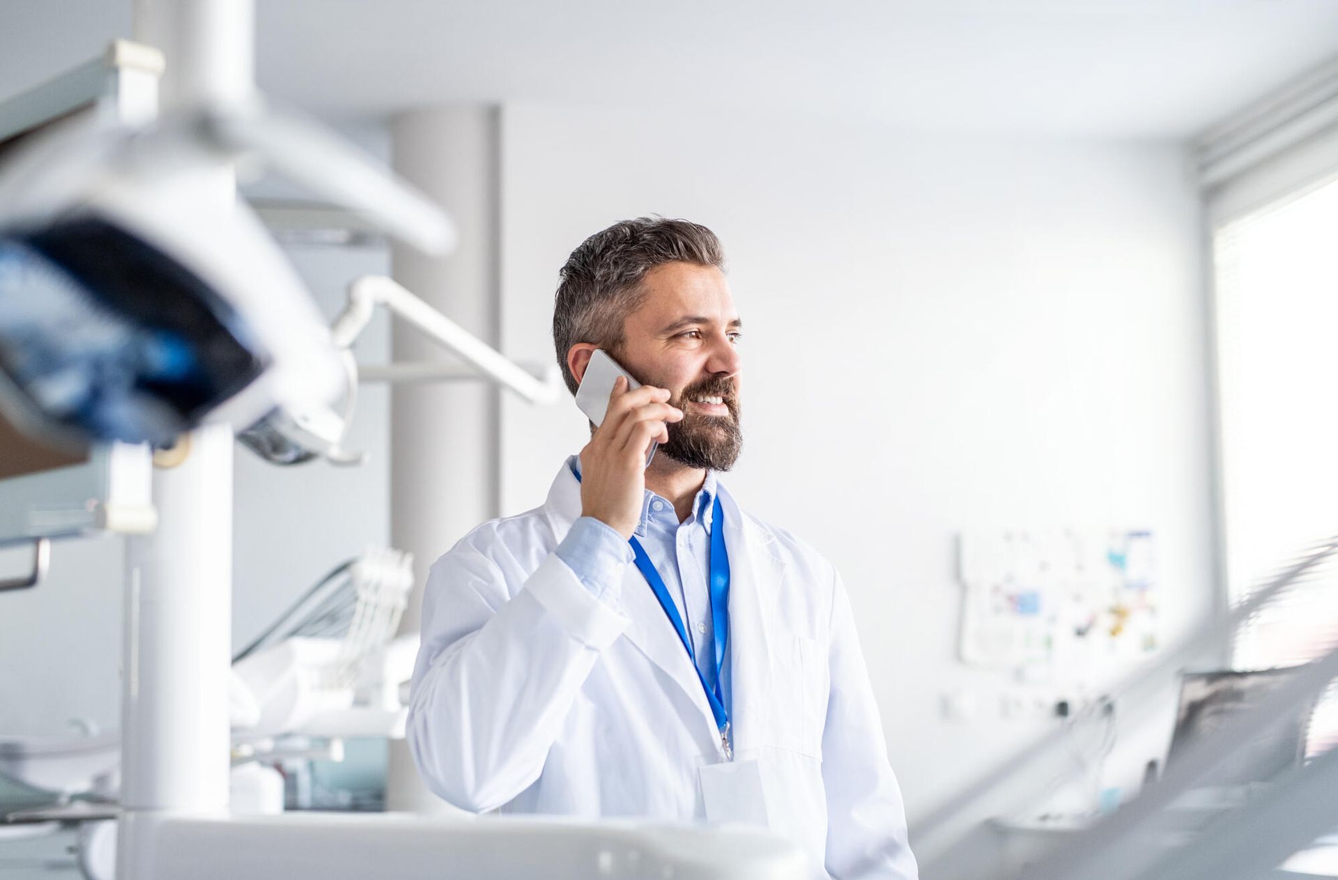 Dentist in white coat smiles while on a phone call in his dental office.