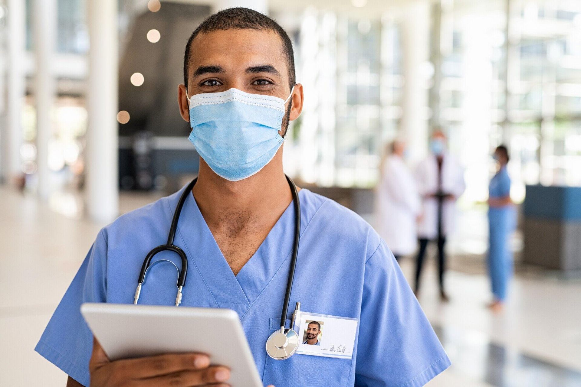Healthcare worker in blue scrubs and mask, holding tablet, stethoscope around neck, hospital background.
