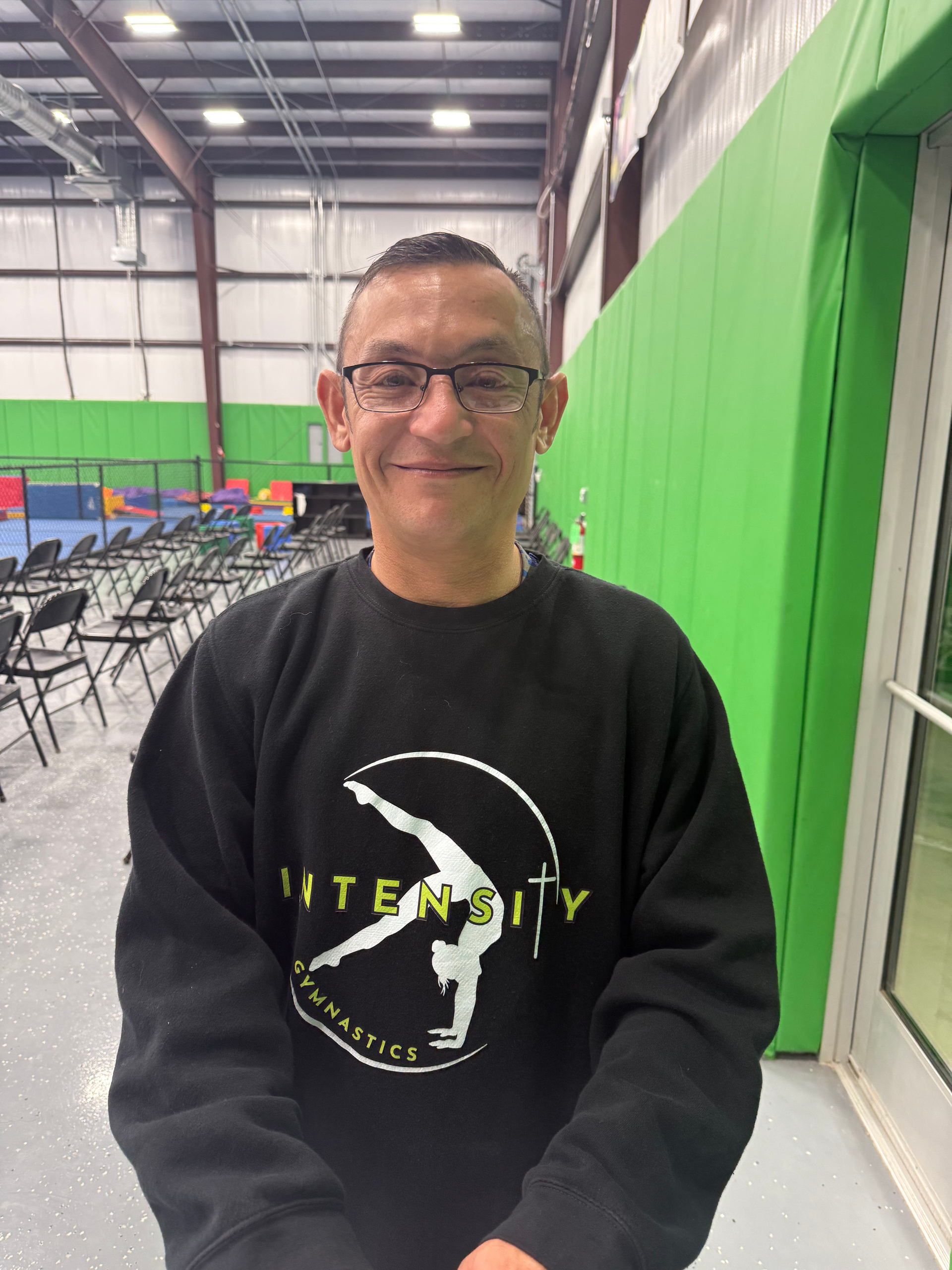 Man in a black sweatshirt with a gymnastics logo, smiling inside a gym.