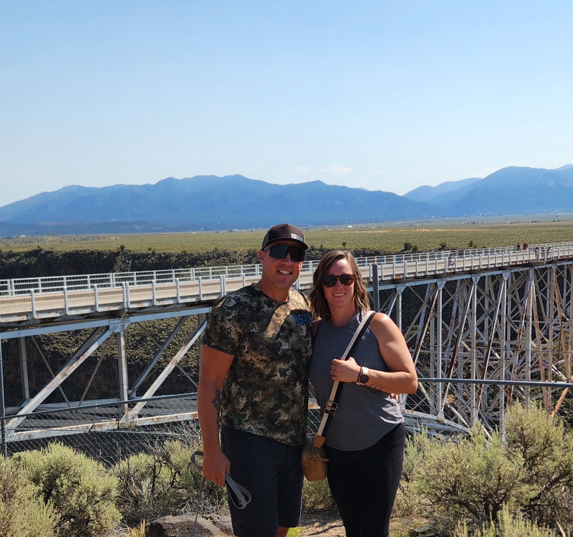 Couple standing near the Rio Grande Gorge Bridge, mountains in background, sunny day.