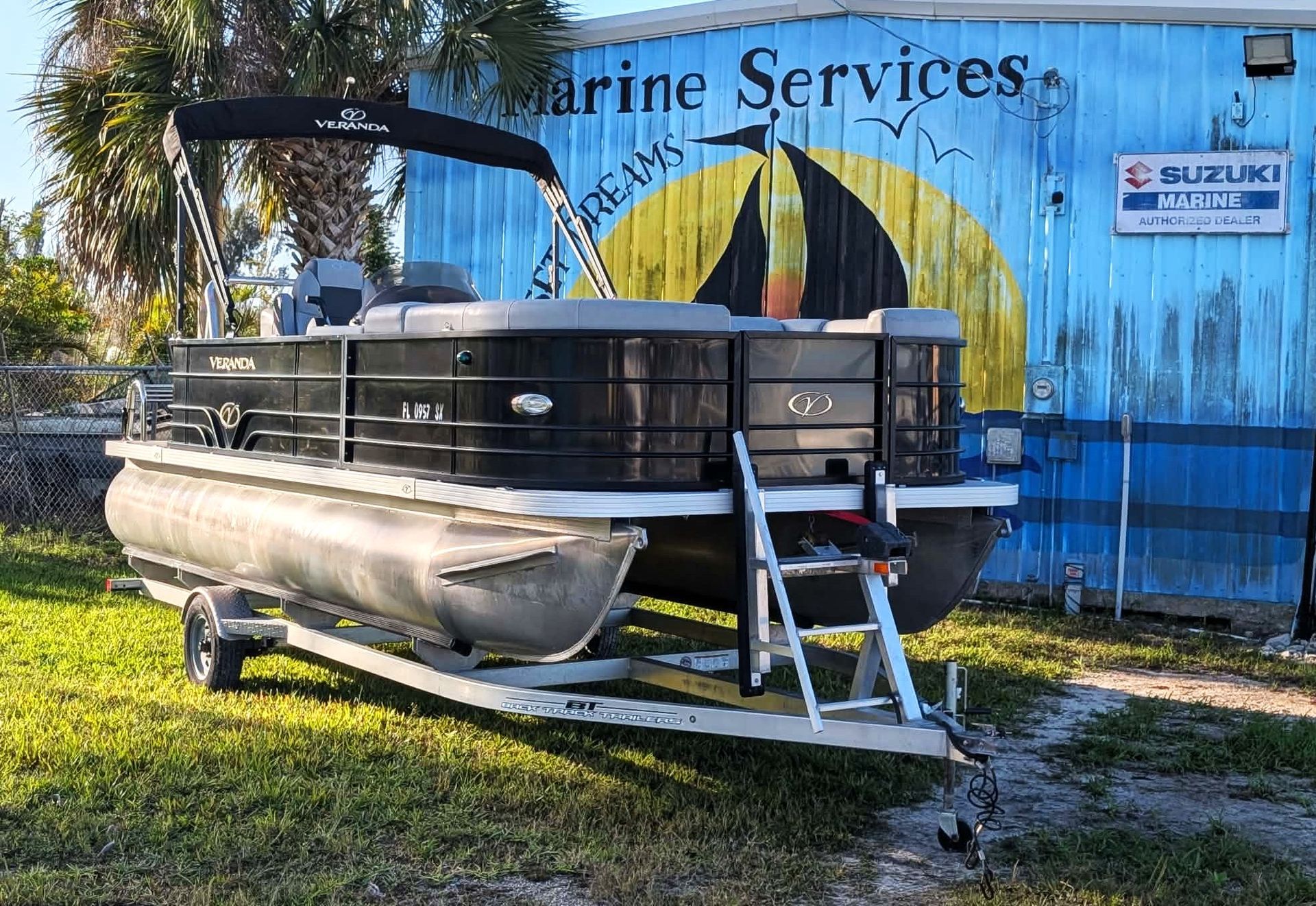 Pontoon boat on a trailer outside a marine services building with a faded mural.