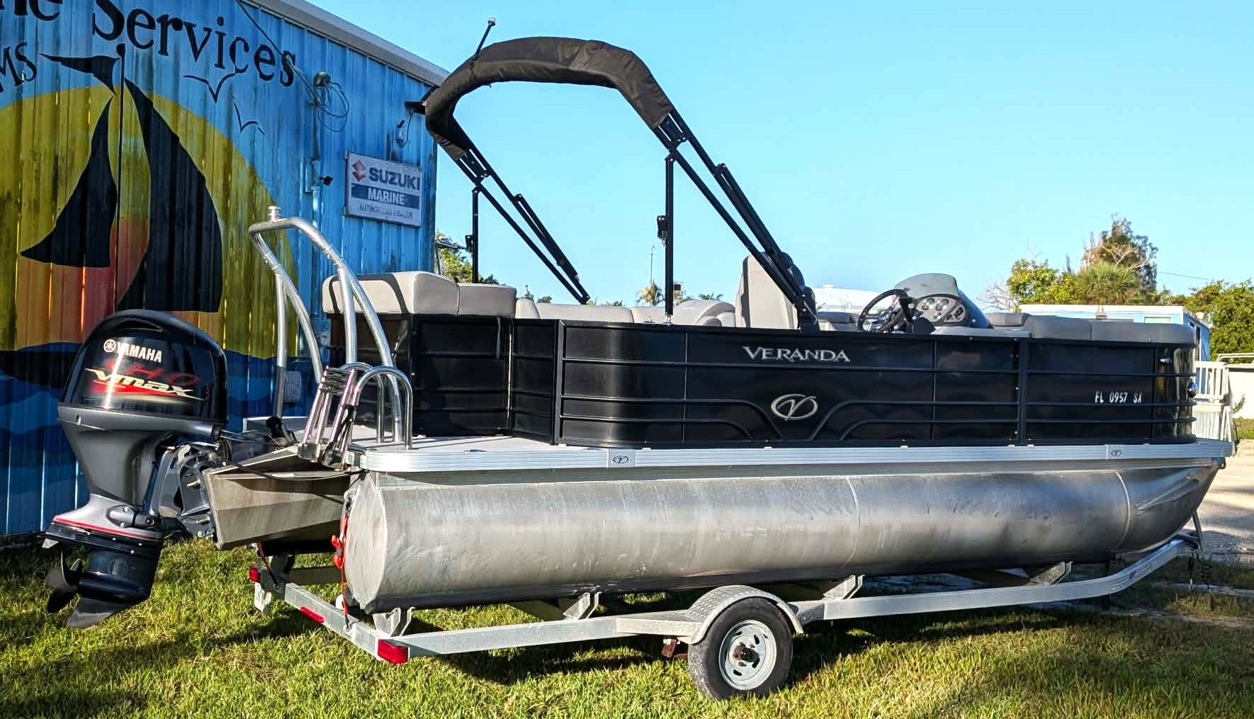 Pontoon boat on a trailer beside a building, with a black canopy and silver pontoons.